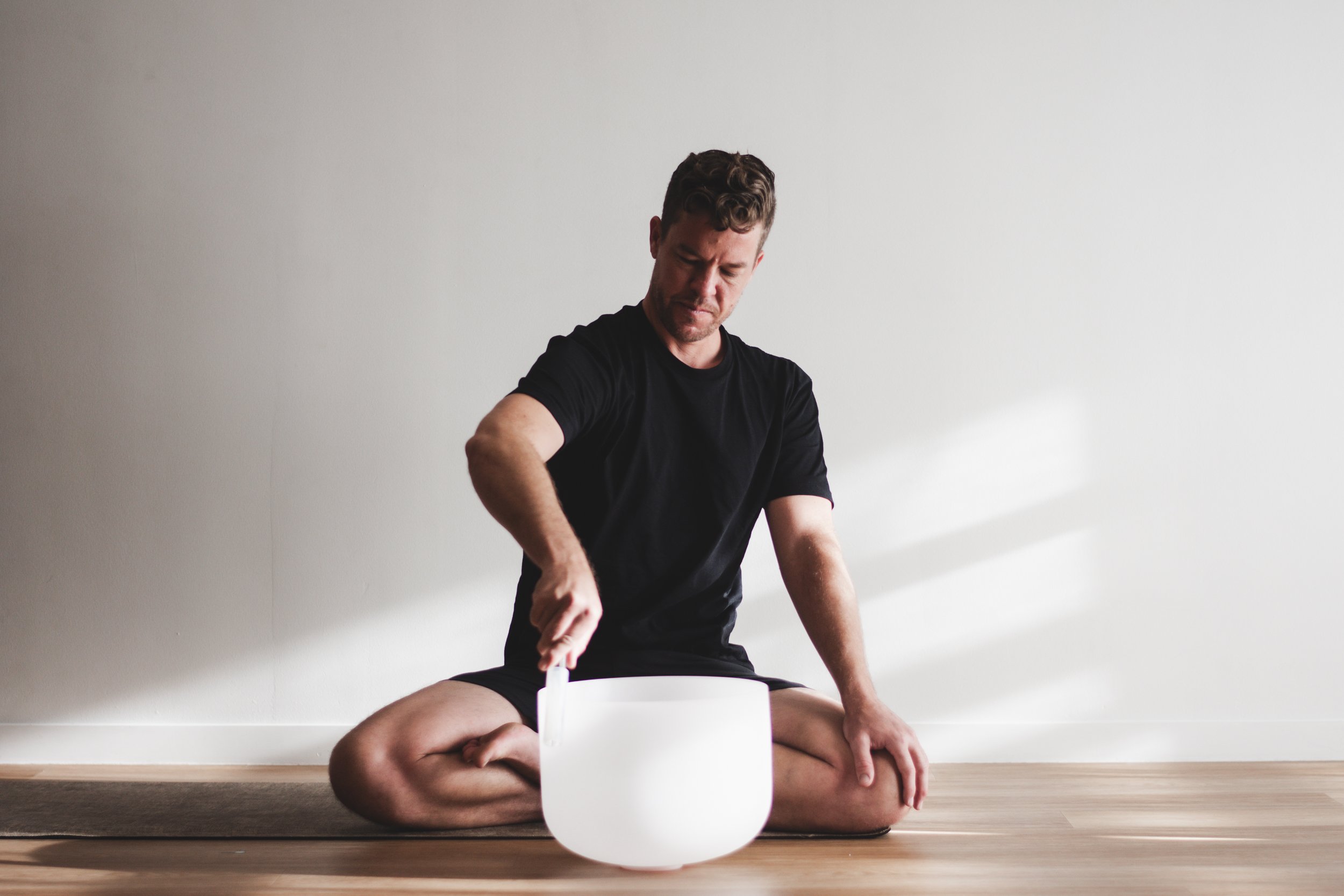 A man sitting cross-legged on the floor, playing a crystal singing bowl for meditation or sound therapy in front of a plain wall.