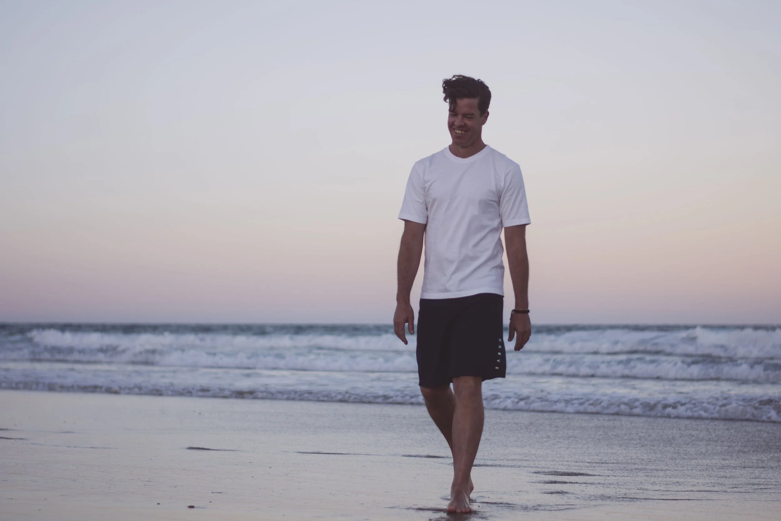 A young man with dark hair, white t-shirt, and black shorts walking barefoot on the beach during sunset, smiling and looking down.