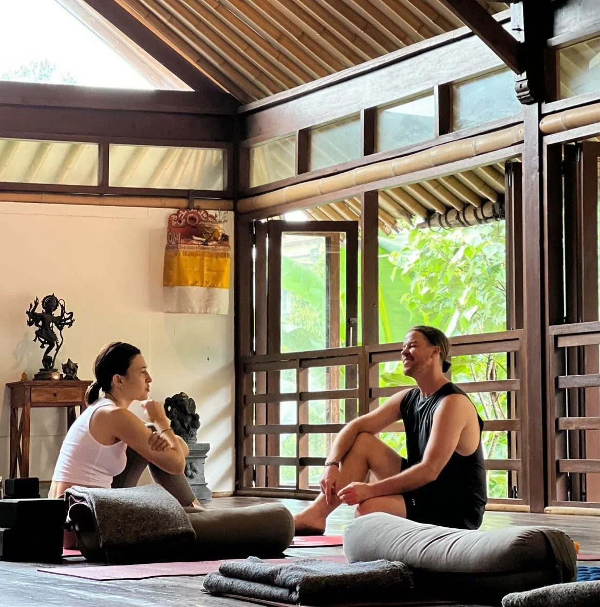 A woman and a man practicing yoga or meditation in a serene, wooden-finished room with large windows showing green foliage outside. They are sitting cross-legged on mats, facing each other and smiling.