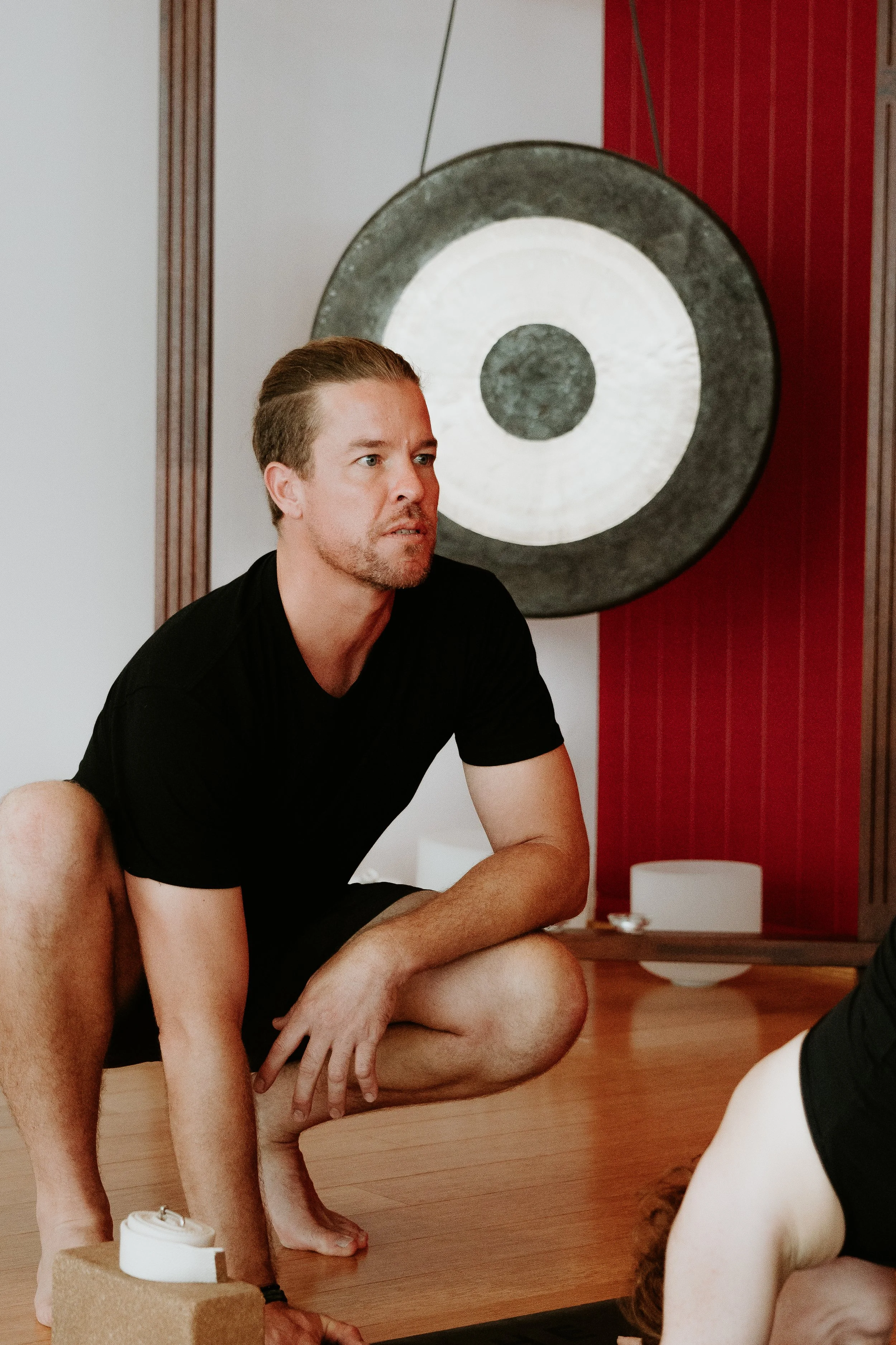 A man with short brown hair, a slight beard, and wearing a black shirt is practicing yoga or meditation in a room with wooden floors and a large gong in the background.