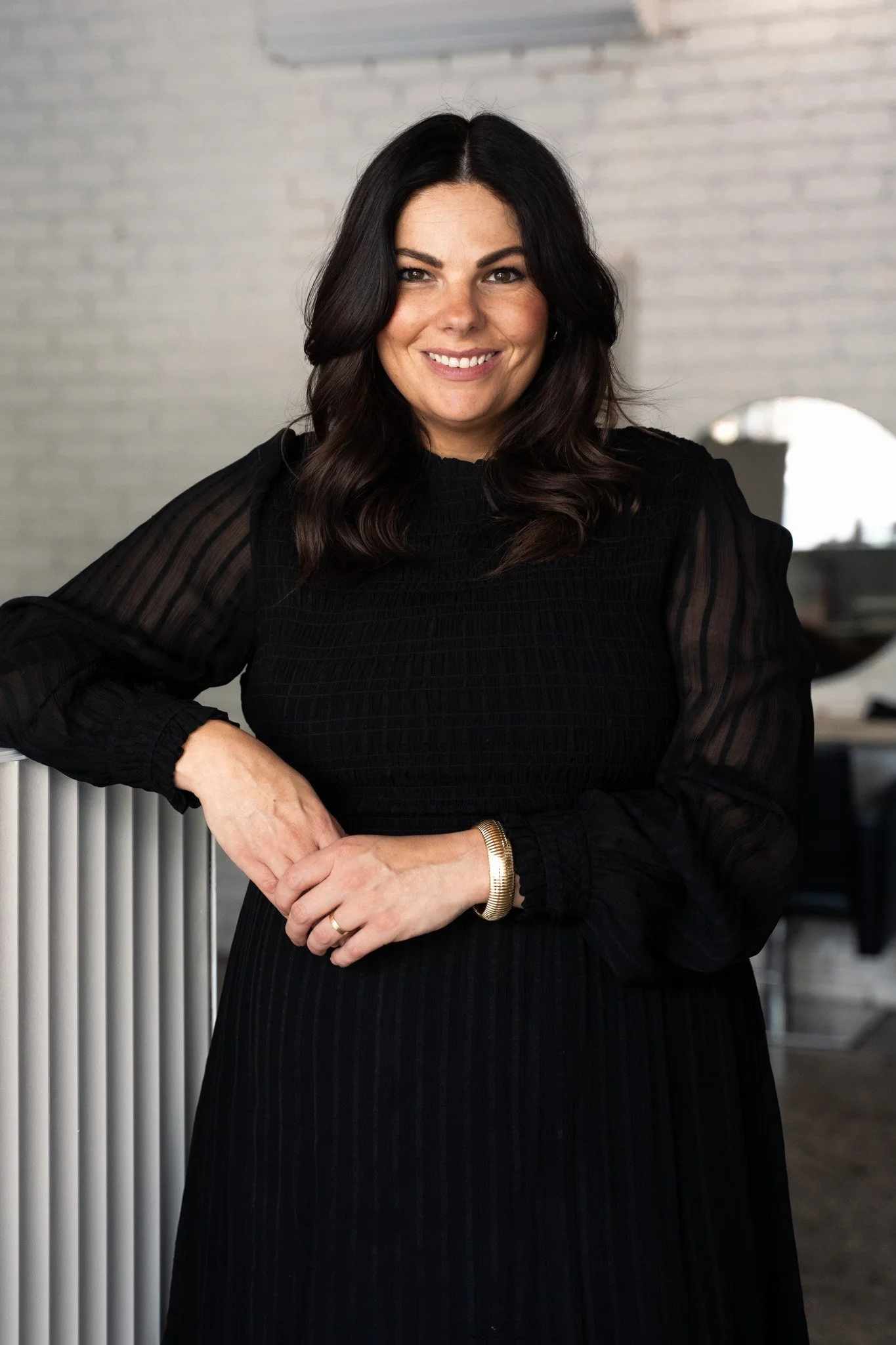 A smiling woman with shoulder-length dark hair, wearing a black blazer, black top, light blue jeans, and black sandals with studded straps, standing indoors near a white wall and sheer curtains.
