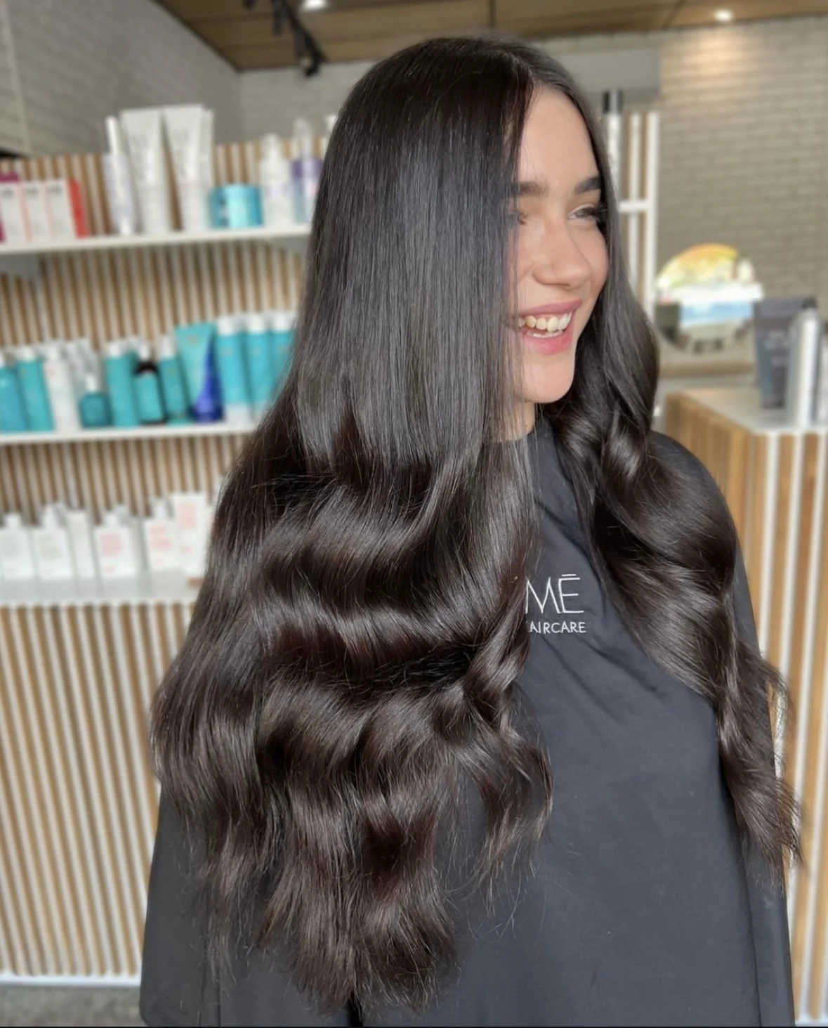 A woman with long, dark, wavy hair smiling in a salon or spa setting.