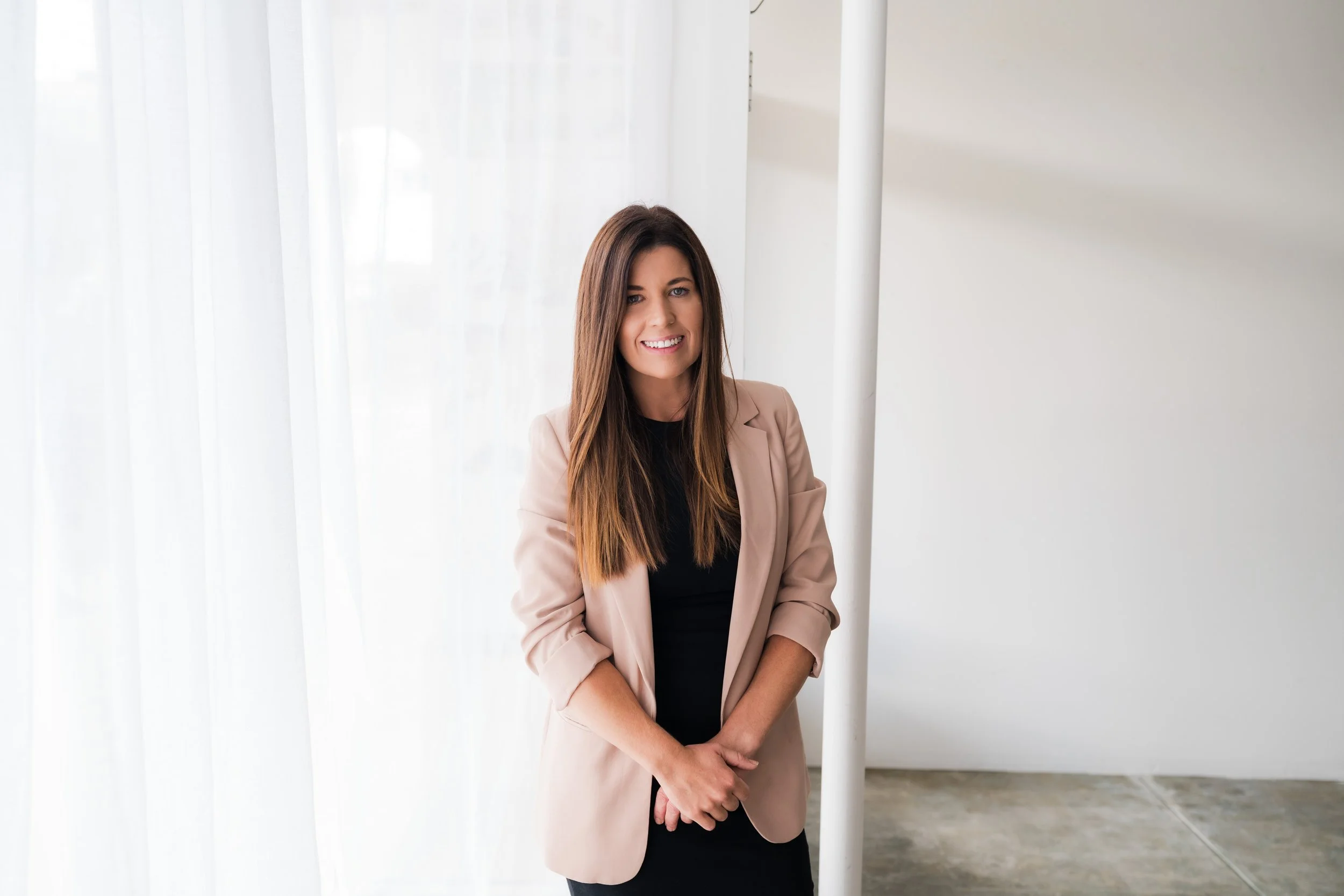 A woman with long brown hair, wearing a beige blazer over a black top, smiling and standing with hands clasped in front of her inside a bright room with white curtains and a white wall.