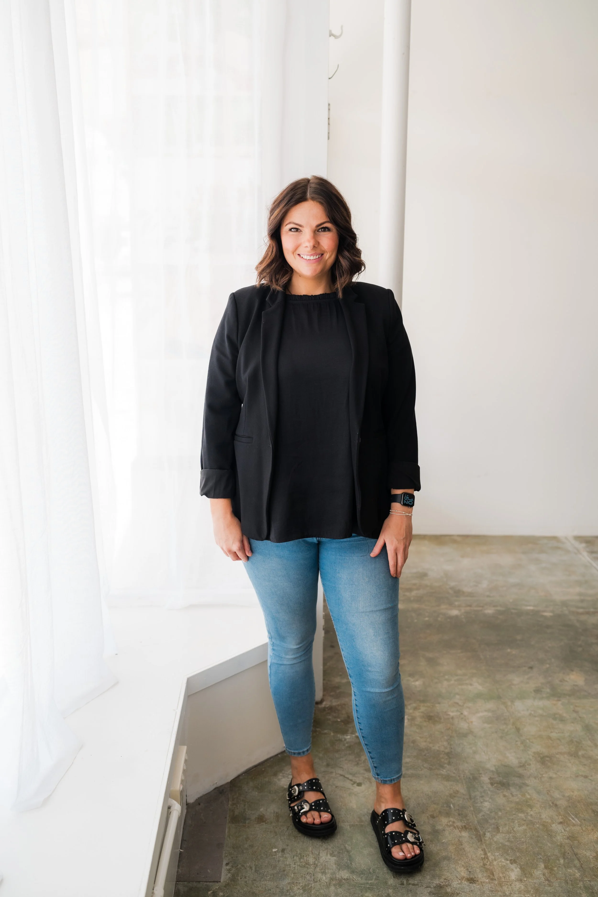 A smiling woman with shoulder-length dark hair, wearing a black blazer, black top, light blue jeans, and black sandals with studded straps, standing indoors near a white wall and sheer curtains.