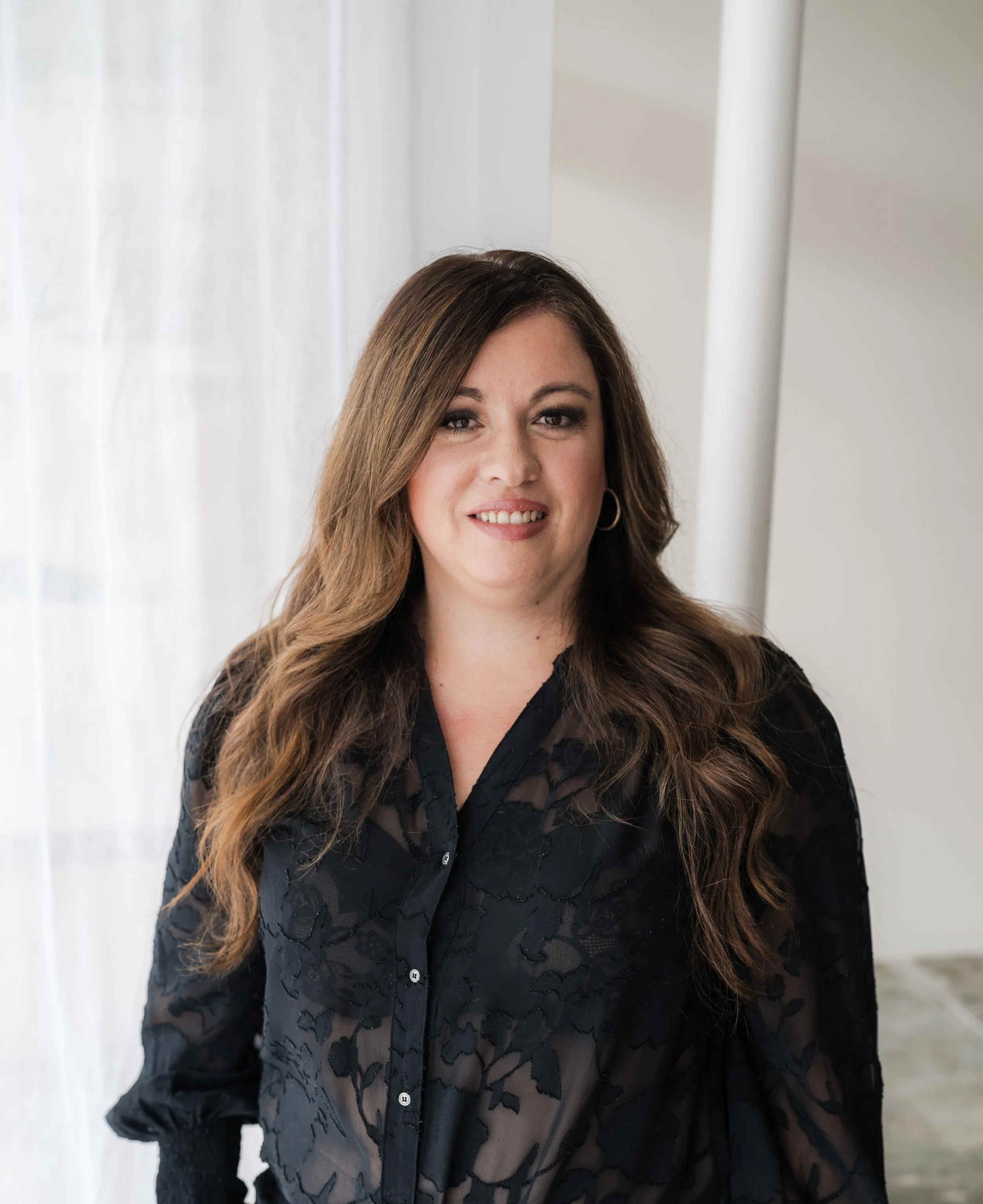 A woman with long brown hair smiling at the camera, wearing a black lace blouse, standing indoors with a white curtain and wall in the background.