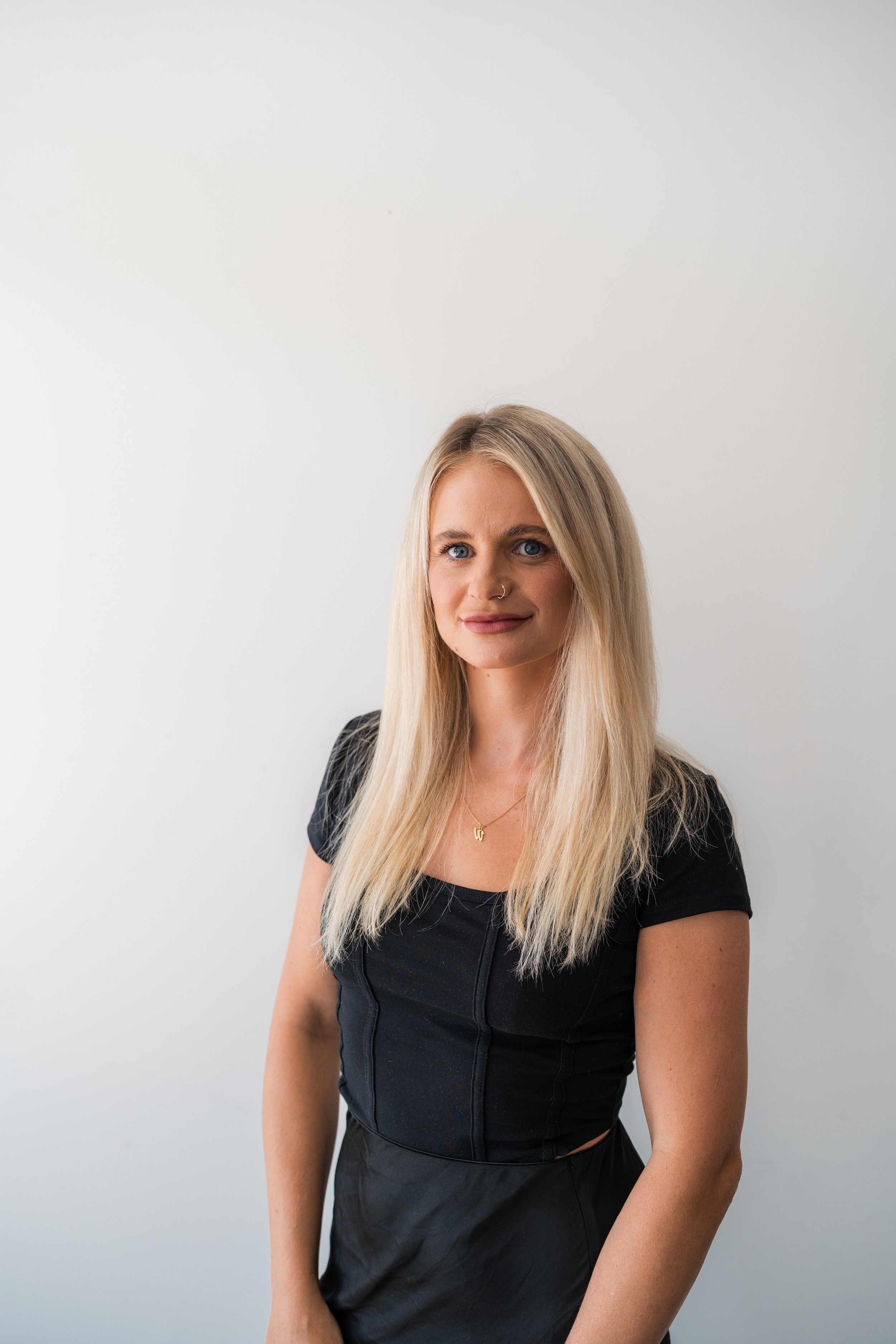 A woman with long blonde hair, blue eyes, and a nose ring, wearing a black top and a gold necklace, standing against a plain white background.