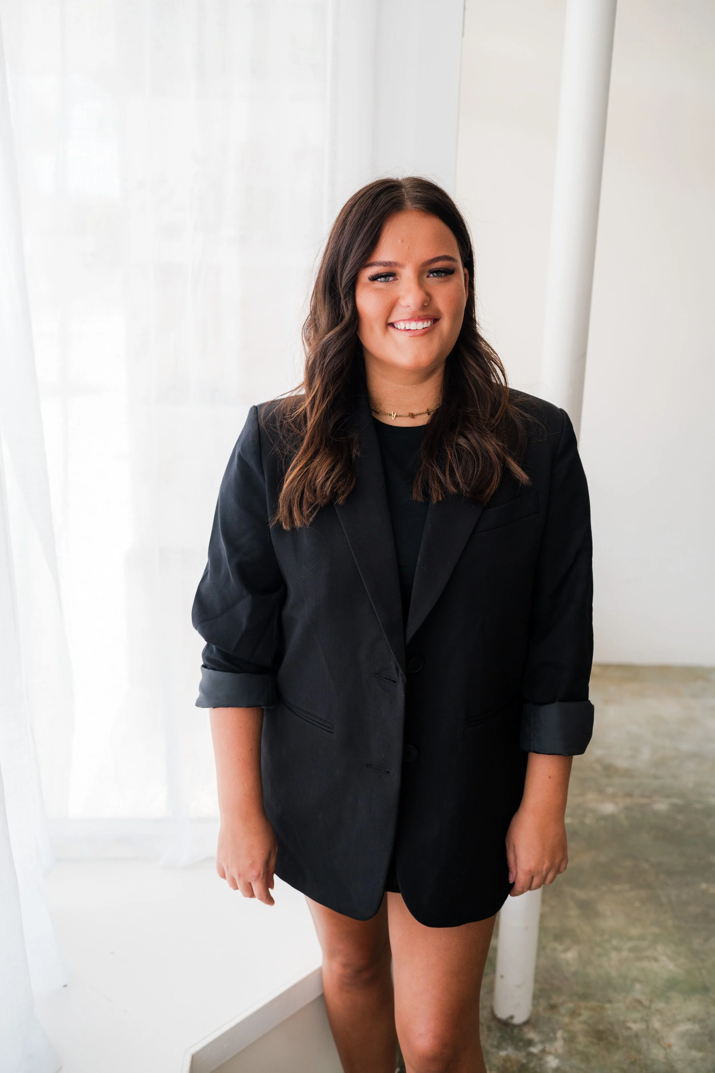 A woman with shoulder-length brown hair, wearing a black blazer and no pants, standing indoors near a window with sheer white curtains.