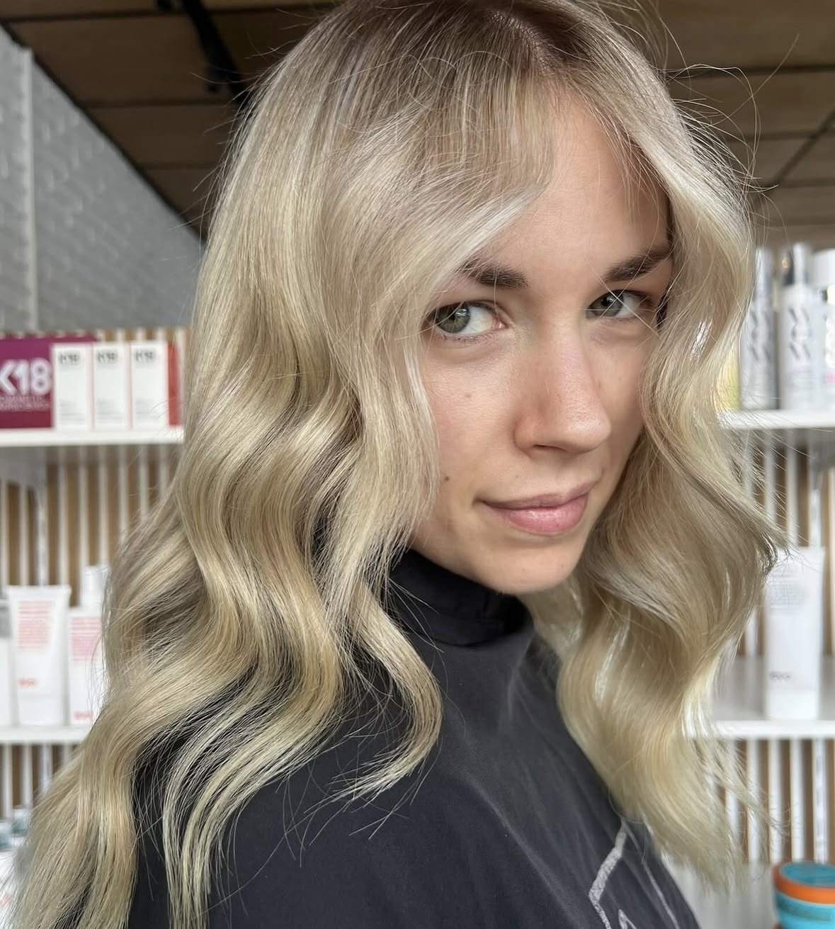 A young woman with long, wavy blonde hair and light skin standing in front of retail shelves with hair care products.