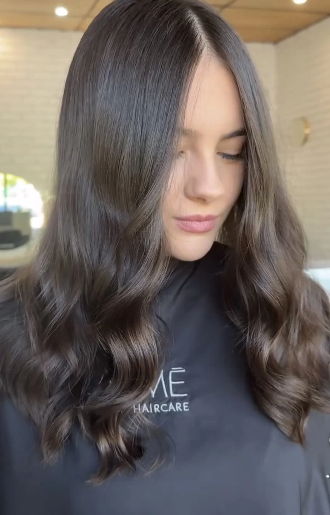 A woman with long, styled, wavy hair is looking downward in a hair salon.