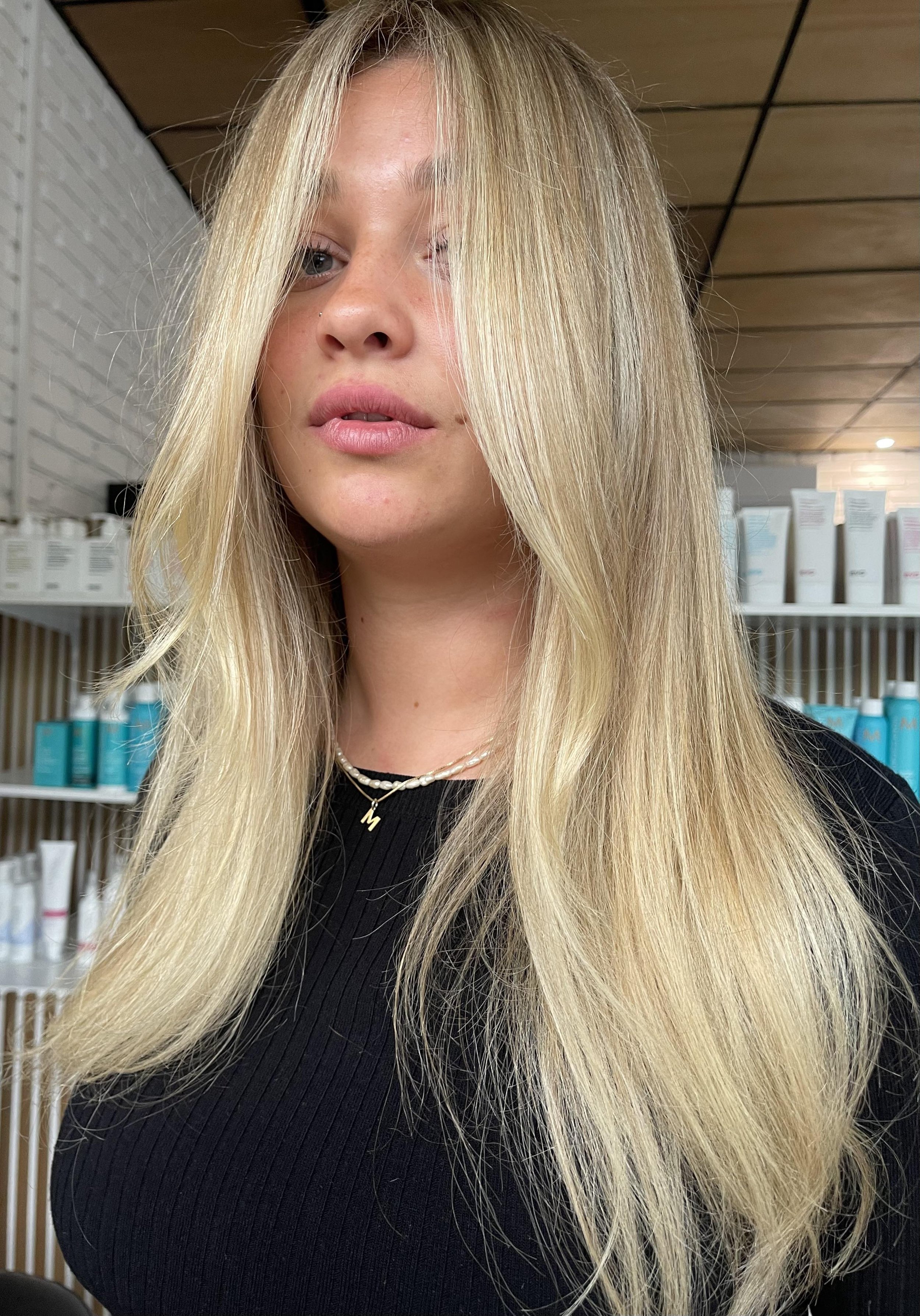 A woman with long blonde hair partially covering one eye, wearing a black top and layered necklaces, standing in front of shelves with beauty products.