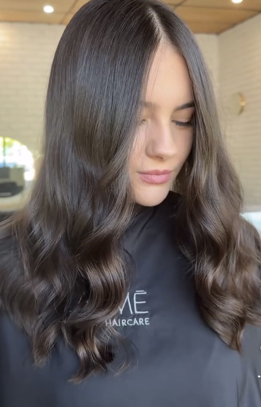 A woman with long, wavy, brunette hair and a light makeup look, wearing a black shirt with the text 'LUME HAIRCARE' on it.