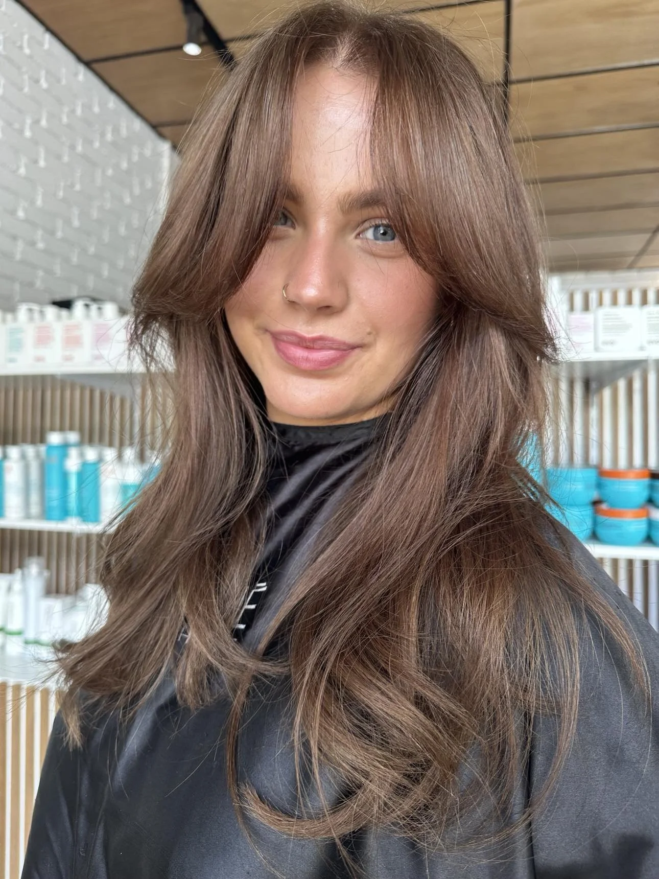 A young woman with long, wavy light brown hair and blue eyes, wearing a nose ring and a black top, standing indoors in front of shelves with hair or skincare products.