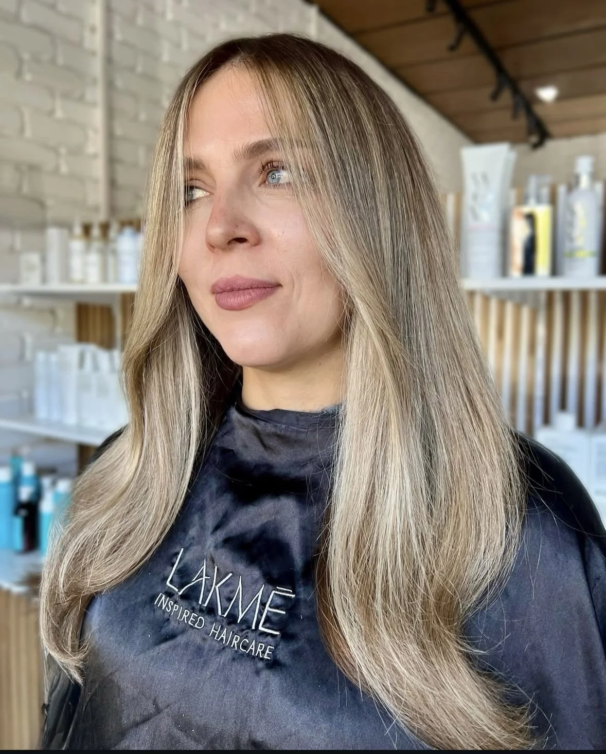 Woman with long blonde hair styled straight, wearing a black salon cape with 'LAKME INSPIRED HAIRCARE' logo, in a salon with shelves of haircare products.
