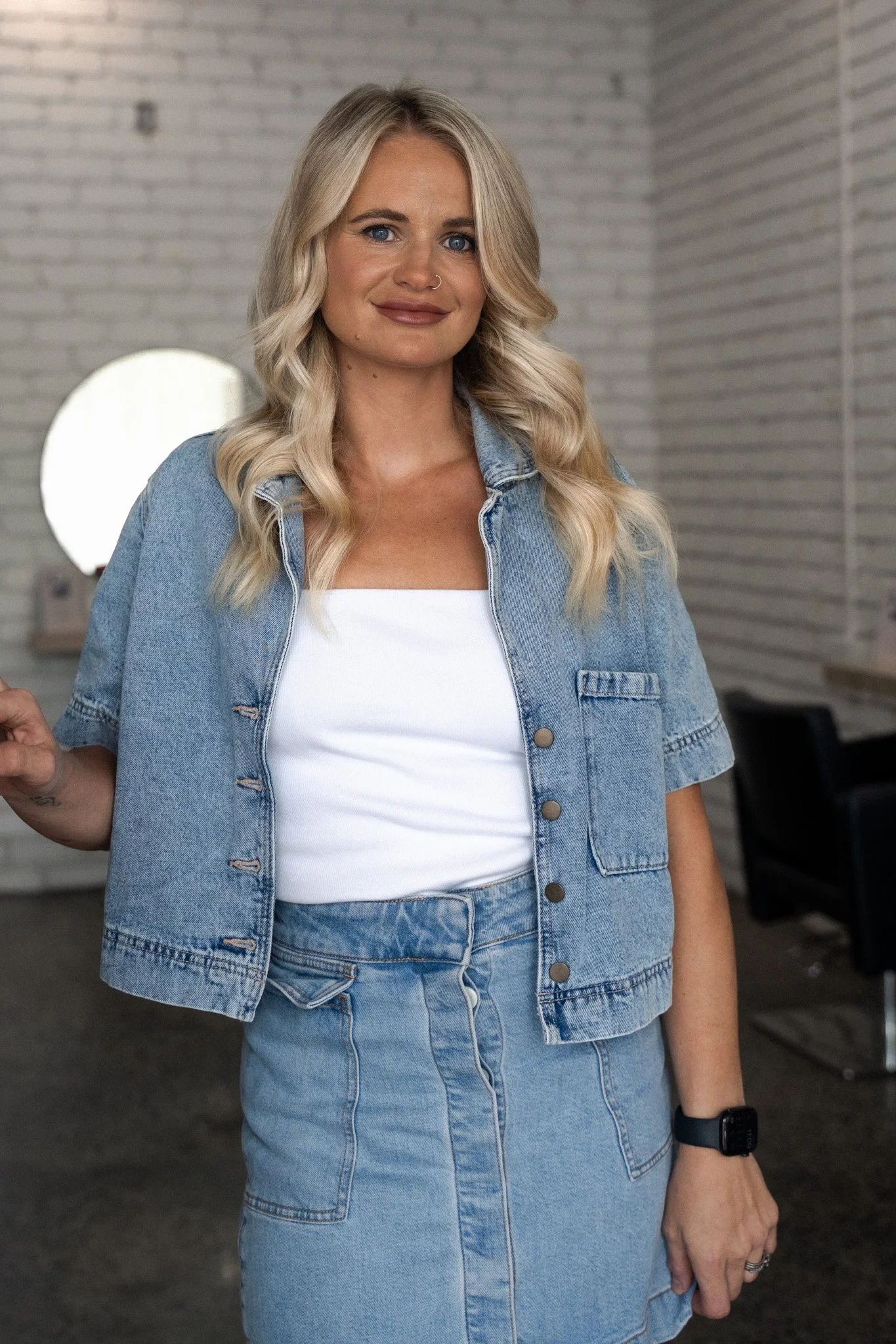 A woman with long blonde hair, blue eyes, and a nose ring, wearing a black top and a gold necklace, standing against a plain white background.