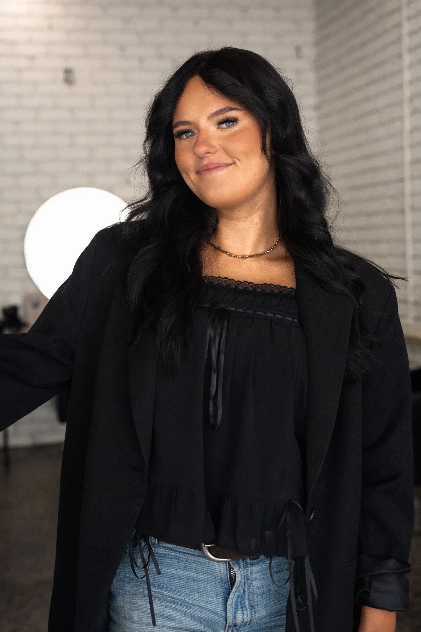 A woman with shoulder-length brown hair, wearing a black blazer and no pants, standing indoors near a window with sheer white curtains.