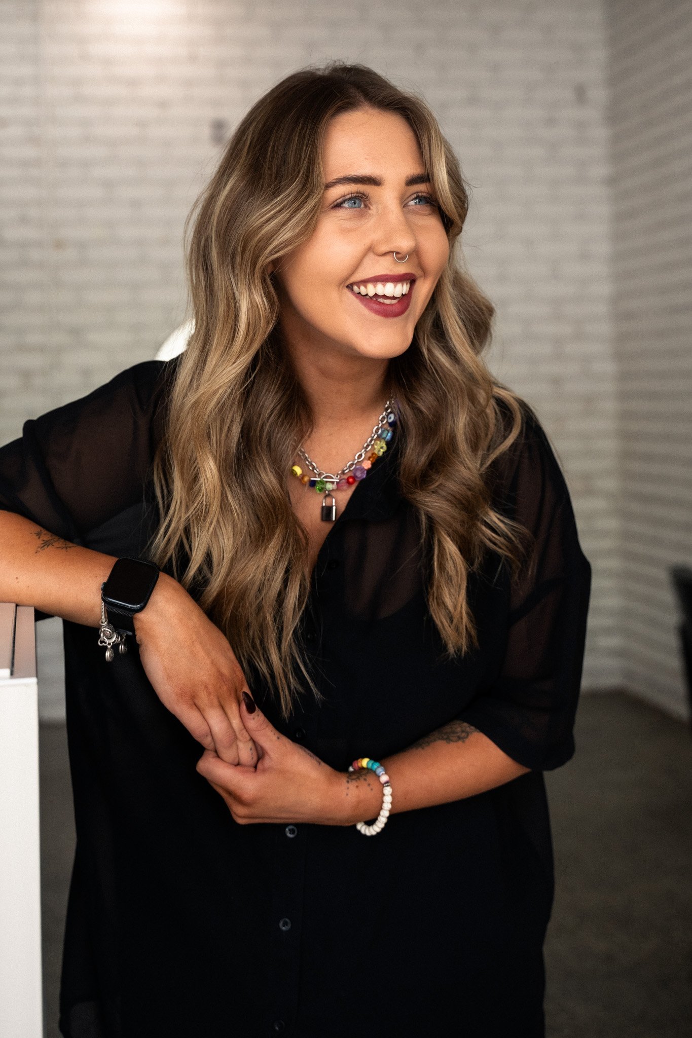A woman with wavy blonde hair, blue eyes, and light skin, wearing a black crop top and black skirt, accessorized with colorful jewelry, standing indoors near a white curtain and wall.