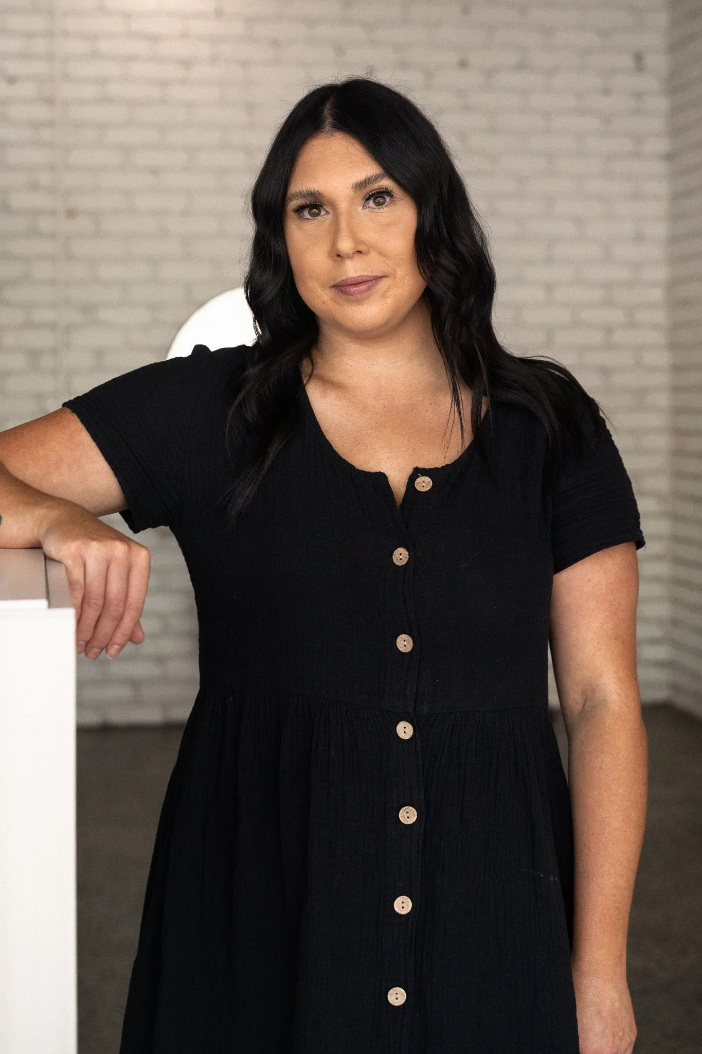 Woman standing indoors near a white wall and curtain, wearing a black dress and black combat boots.