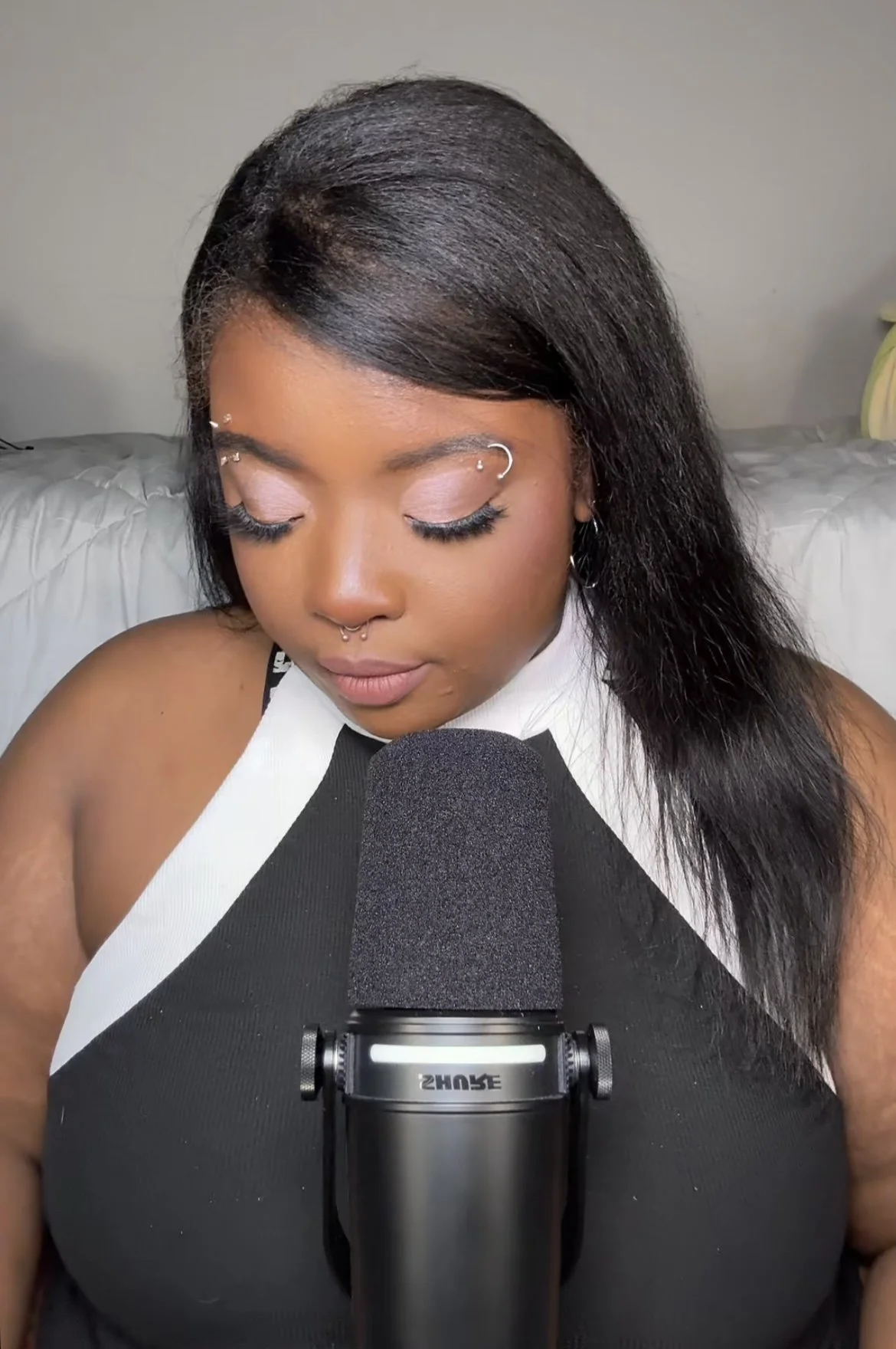 A woman with long black hair and piercings, wearing makeup with eyes closed, sitting in front of a microphone in a room with gray walls and a bed in the background.