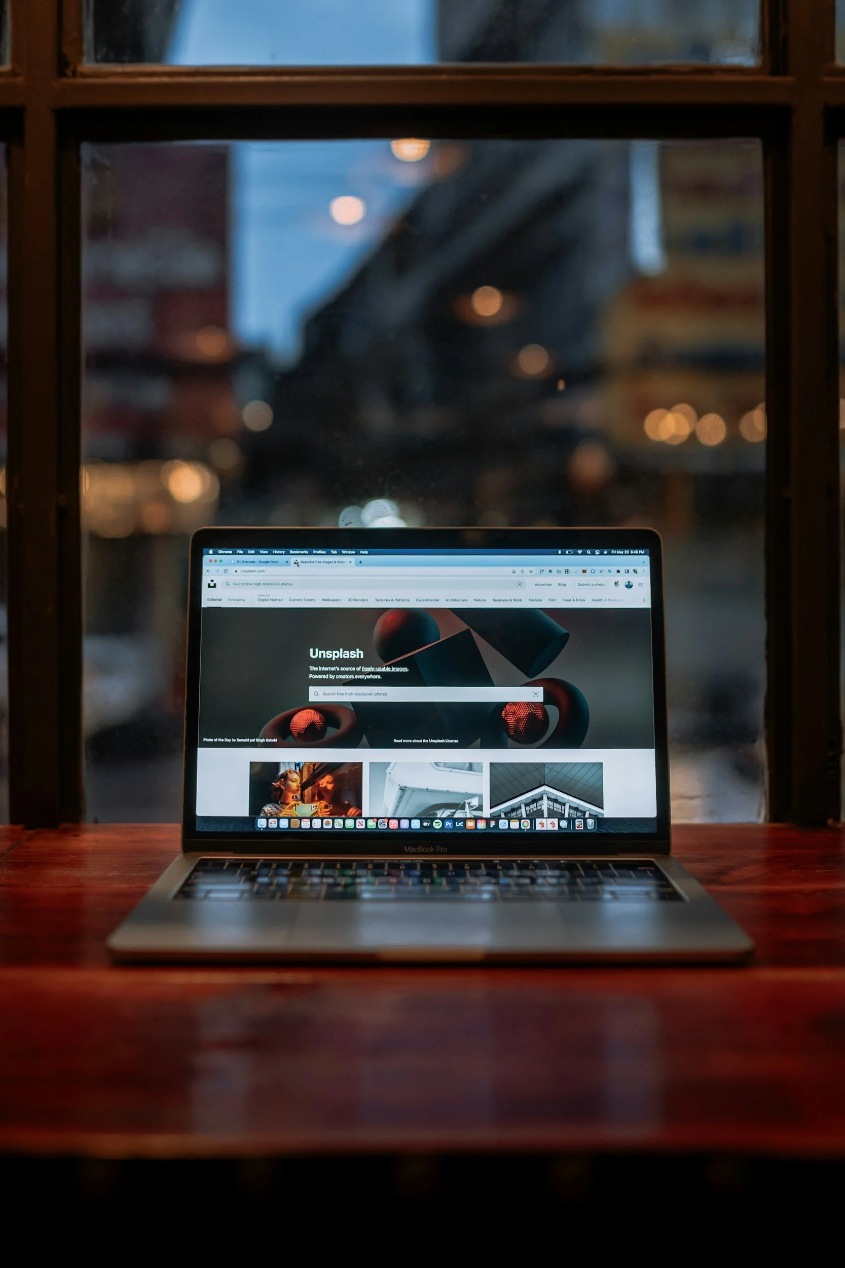 Open MacBook Pro displaying the Unsplash website on a wooden table, with blurred city lights and buildings visible through the window behind.