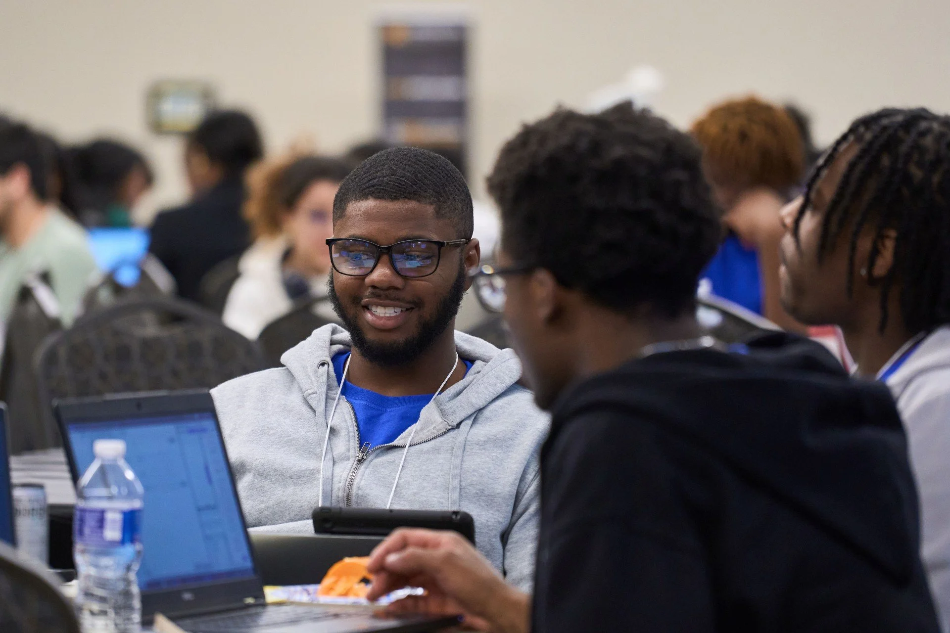 Three young men are sitting at a table during a conference or workshop. The man in the middle is smiling, wearing glasses, a gray hoodie, and blue shirt, and looking at two men in front of him, who are engaged in conversation. There are laptops, a water bottle, and snacks on the table.
