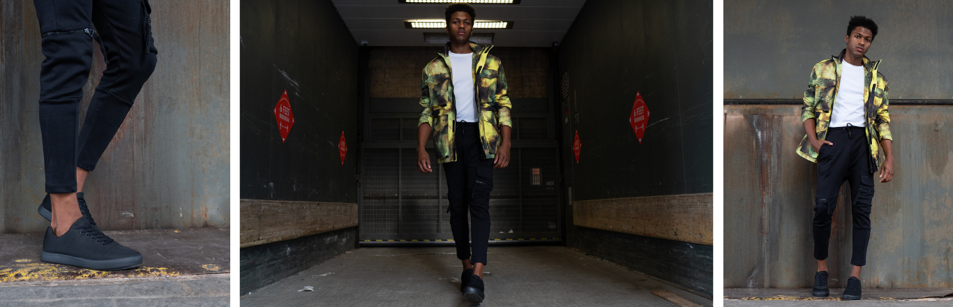 A young man walking in an urban setting, wearing a camo jacket, white t-shirt, black pants, and black shoes.