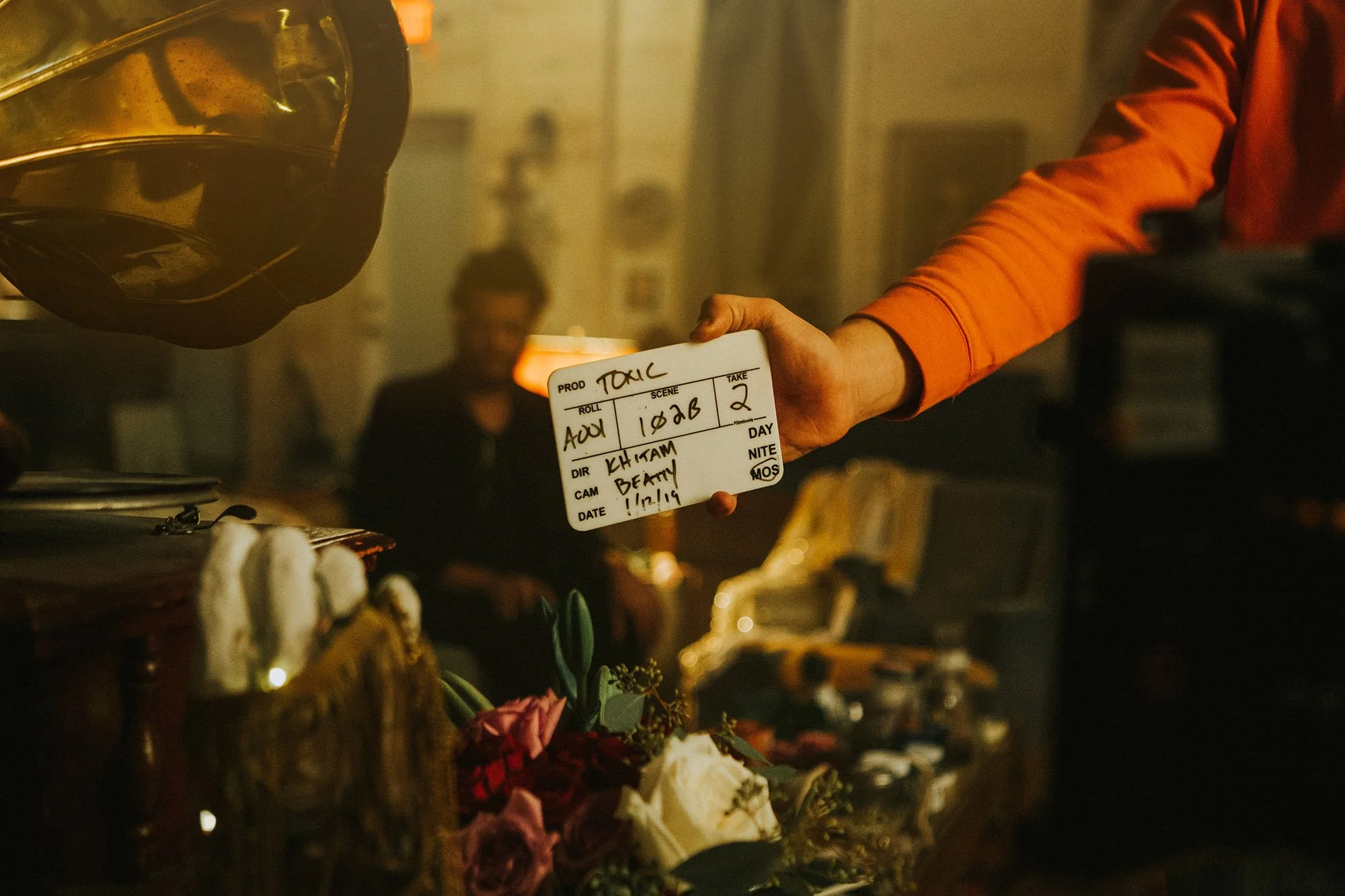 A person holding a clapperboard on a film set, with a woman sitting in the background, and a table with flowers in the foreground.