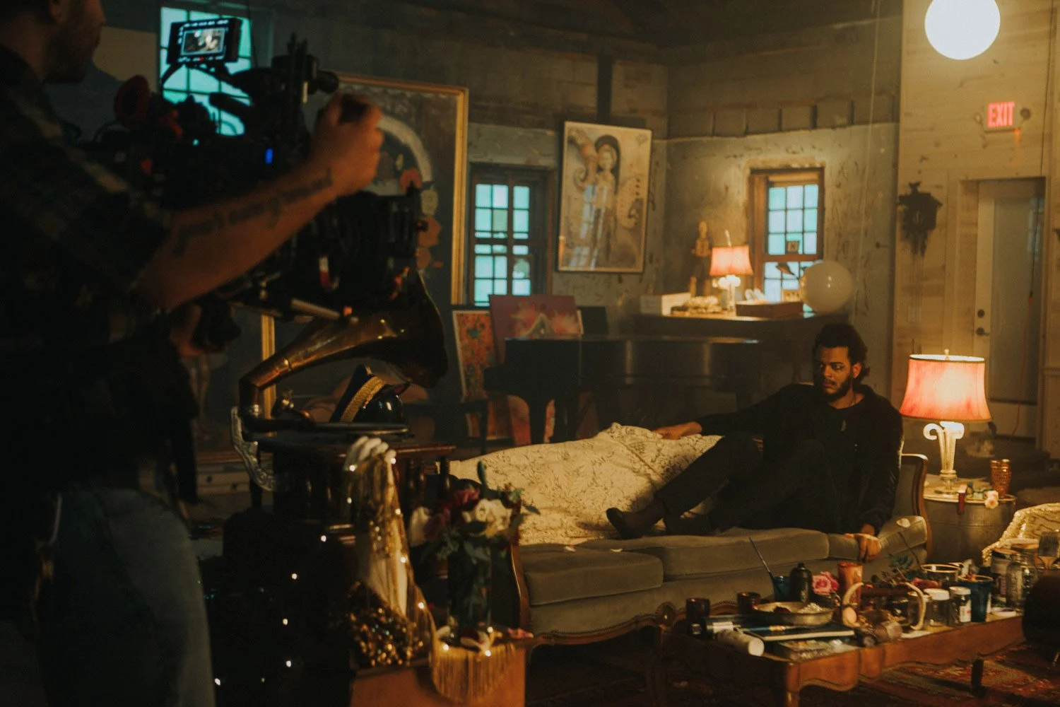 Man lying on vintage sofa in dimly lit room with eclectic decor, paintings, and lamps, while a person operates camera nearby.