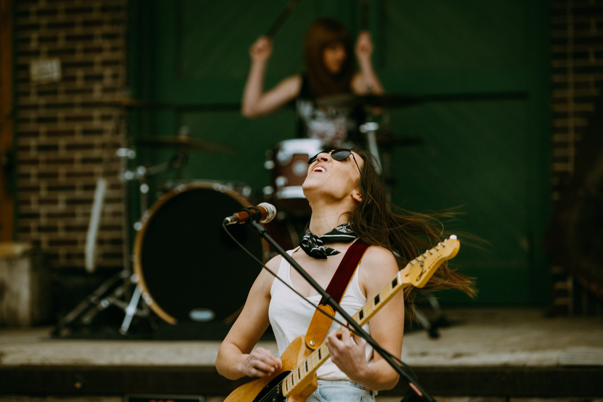 A female musician passionately playing an electric guitar and singing into a microphone with her eyes closed, wearing sunglasses and a bandana. A drummer is in the background playing drums on a stage.