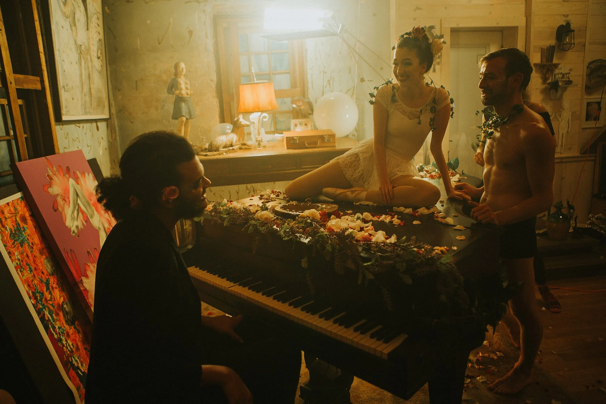 Four people in an art studio, two nude and two clothed, engaging in a playful, intimate scene around a decorated piano.