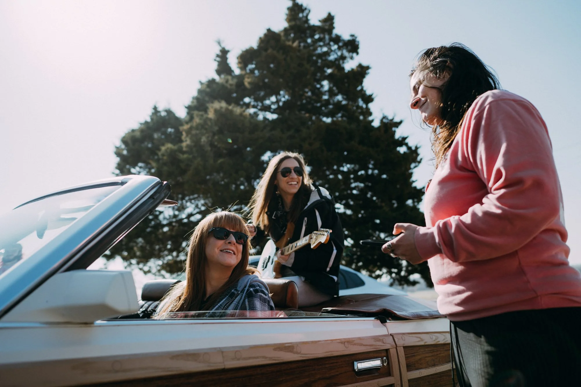 Three women enjoying a sunny day outdoors near a vintage convertible car, with one woman sitting inside the car and two women standing outside, one of whom is playing guitar.