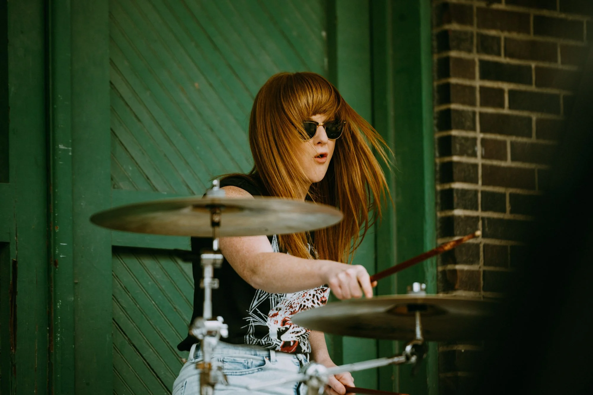A woman with long red hair wearing sunglasses is playing drums outdoors near a green door and brick wall.