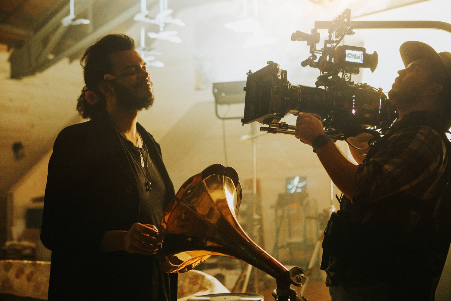 A woman holding a vintage gramophone being filmed by a cameraman with a professional camera in a dimly lit indoor setting.