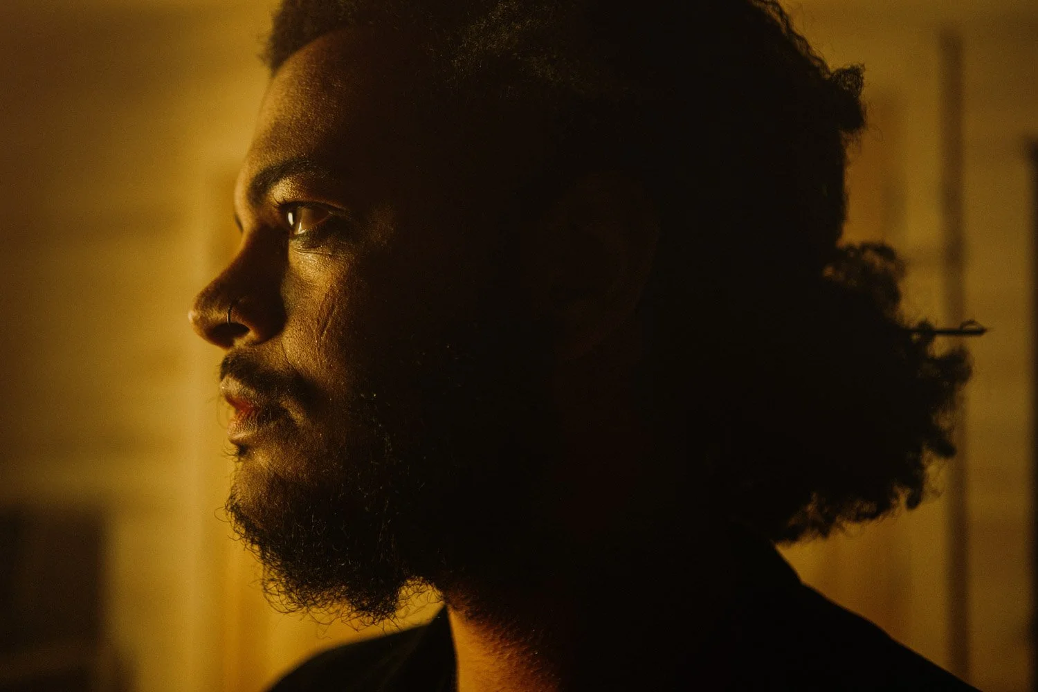 A side profile of a man with dark skin and curly hair, illuminated by warm, low lighting.