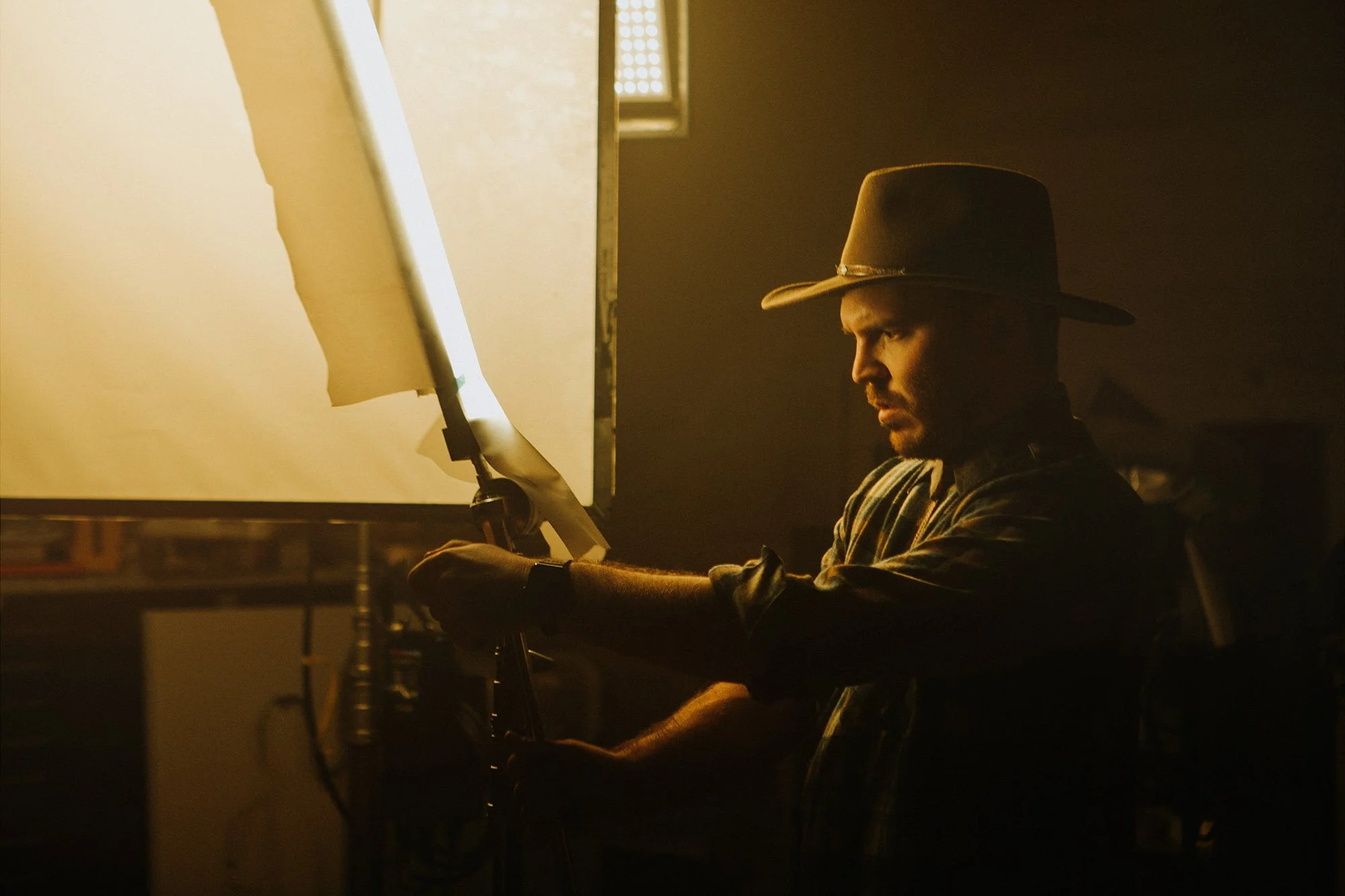 A man wearing a cowboy hat working on film set with a light reflector in a dimly lit room.