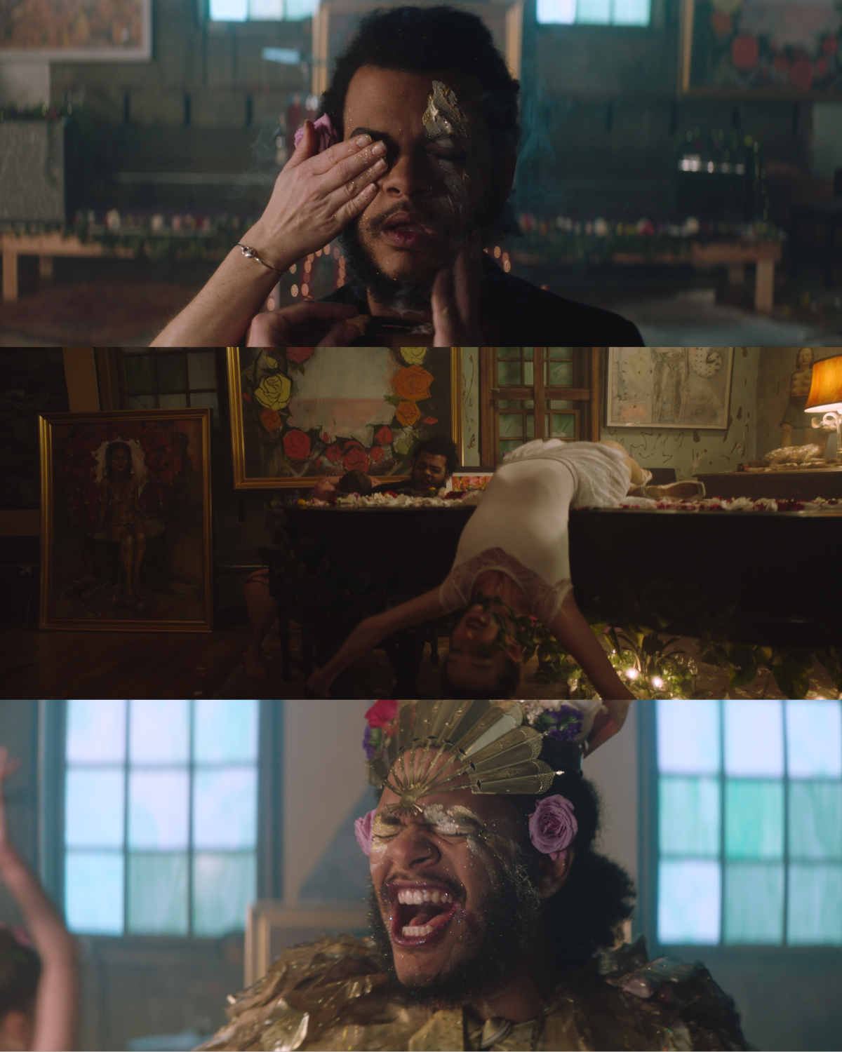 Three scenes show a man with a beard and tattoos, involved in artistic, colorful activities. In the first scene, he wipes his face with a cloth, in the second scene, he is lying on a table covered in flowers with a woman upside-down behind him, and in the third scene, he wears an ornate headpiece and gold costume, smiling with paint and glitter on his face in a well-lit room.
