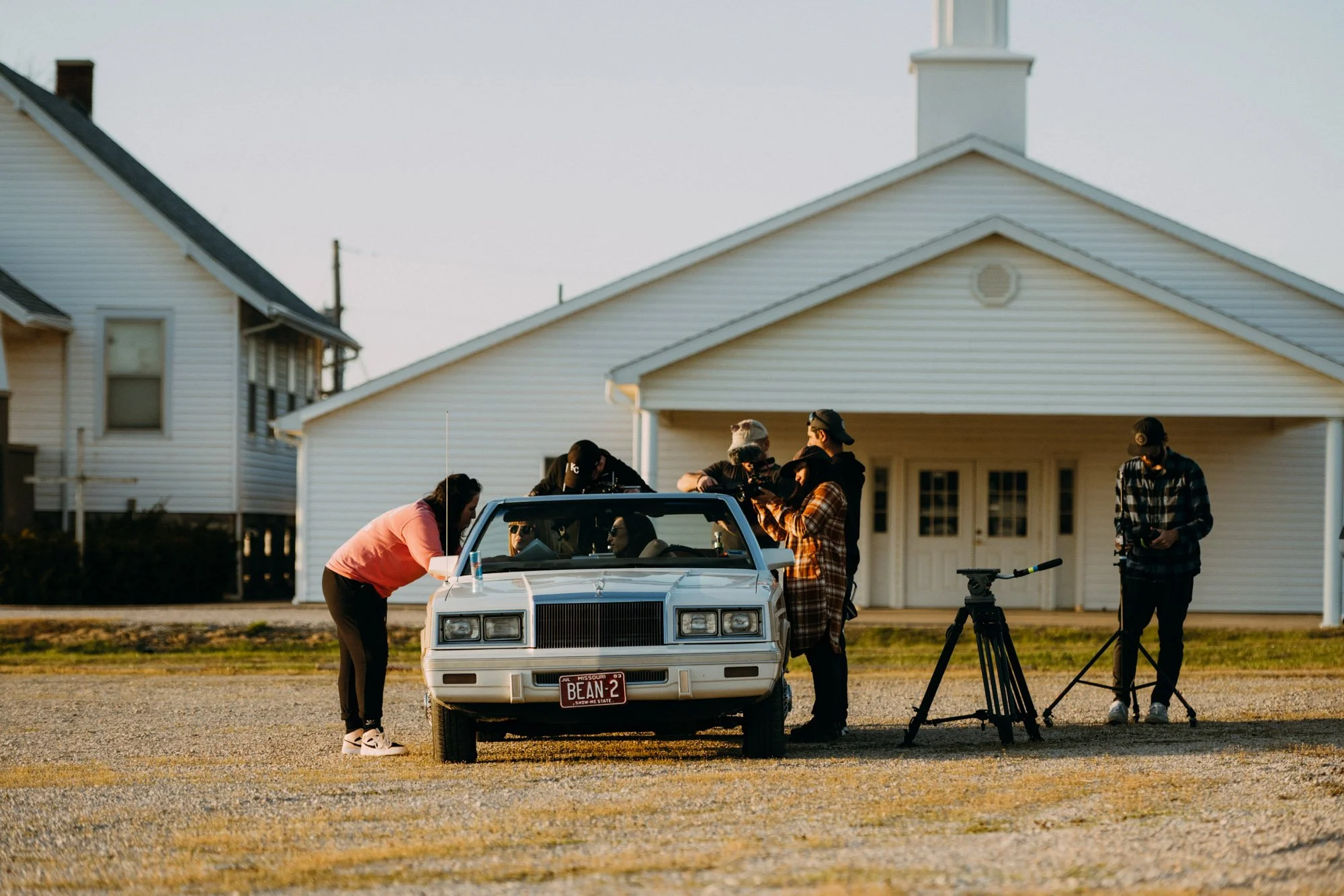 A film crew shooting a scene outdoors next to a white car with a 'Missouri Bean' license plate, in front of a white house during sunset.
