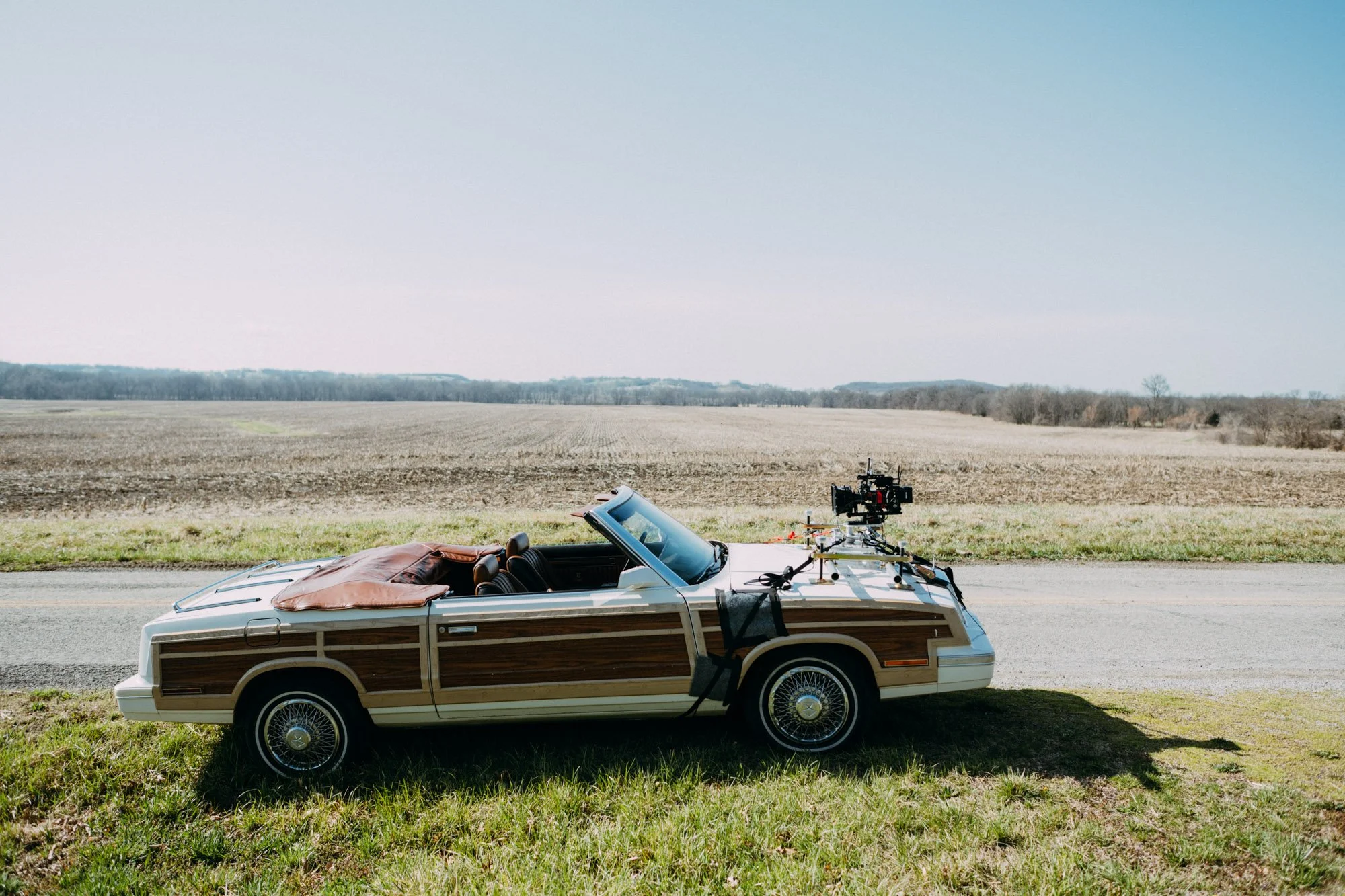 A vintage station wagon with a wood panel exterior parked on grass beside a road in a rural area, equipped with filming equipment on the roof, overlooking open fields and a clear sky.