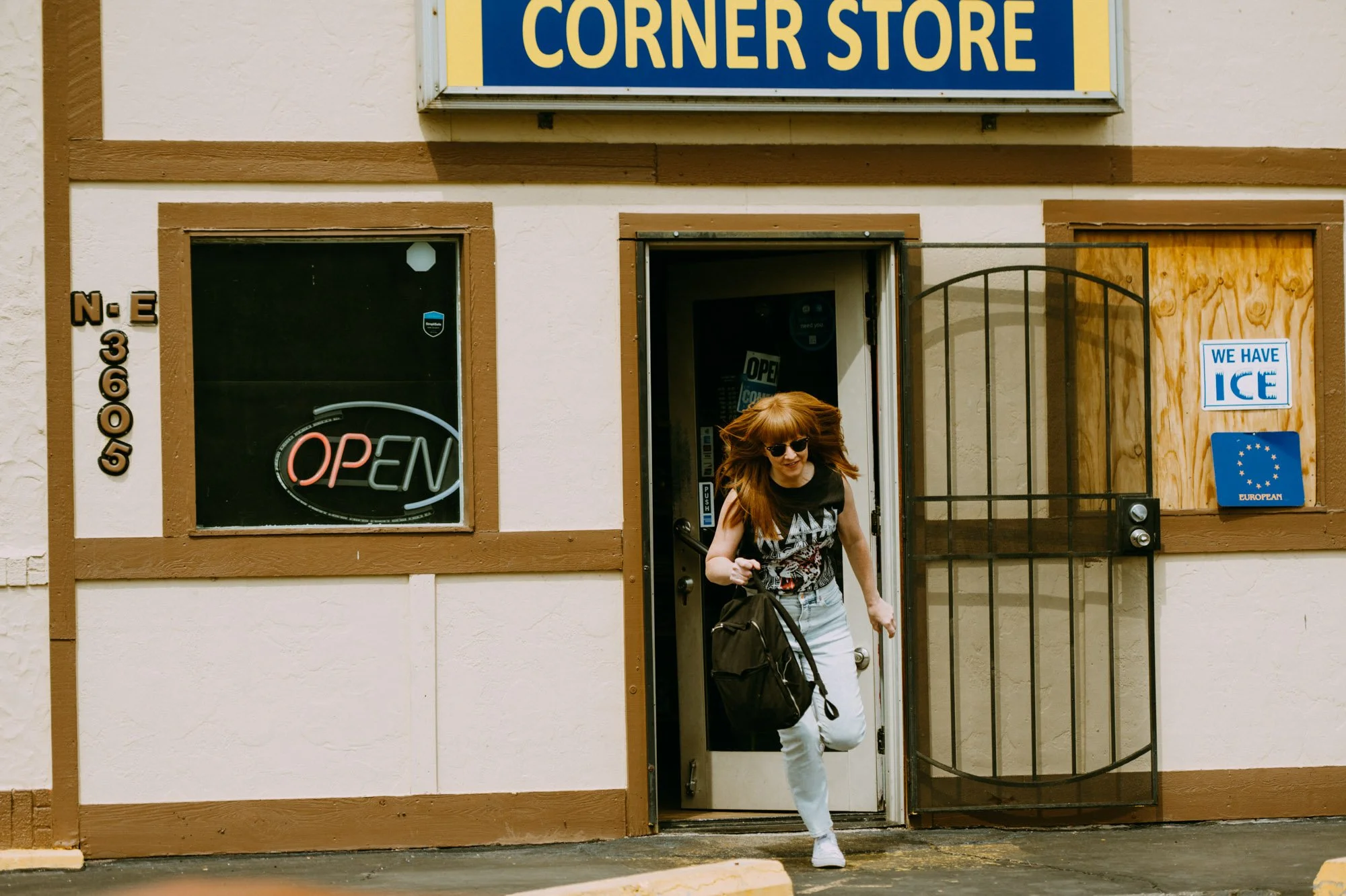 A woman with red hair, sunglasses, and a black T-shirt running out of a convenience store with a black backpack.