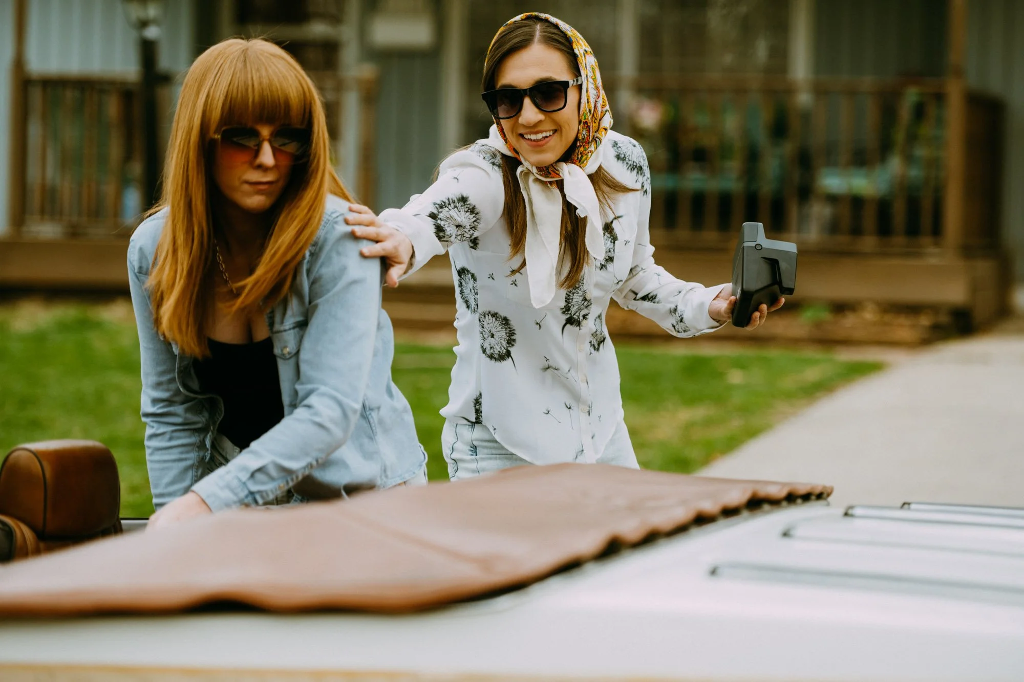 Two women outdoors, one with red hair wearing sunglasses and a denim jacket, the other with brown hair, a floral headscarf, and sunglasses, smiling and touching the shoulder of the first woman, with a large brown bag and a table or surface in the foreground.