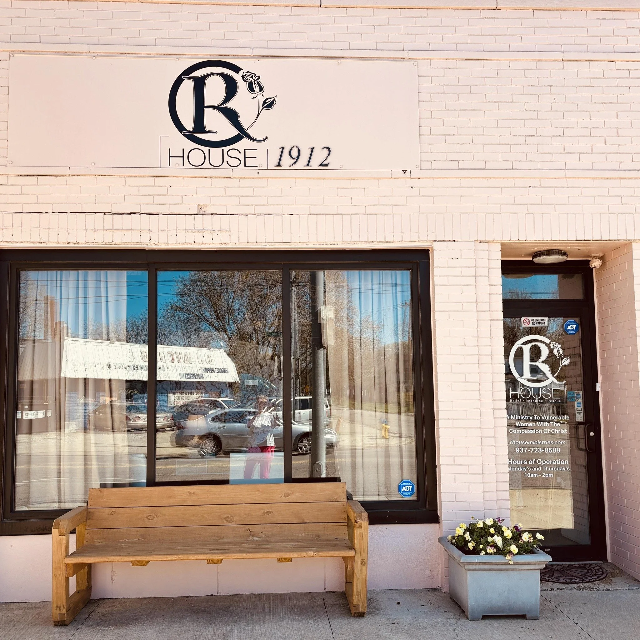 Front of a pink brick building with a sign that reads 'R House 1912' and a logo of a letter R with a rose. There is a glass door and a large window, with a wooden bench and a flower planter outside.