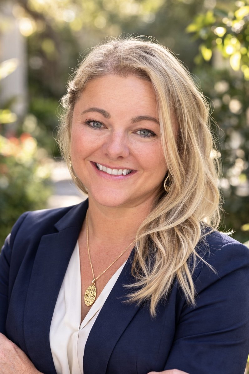 A woman with blonde hair and a maroon top taking a selfie on a wooden bridge overlooking a river with autumn foliage.