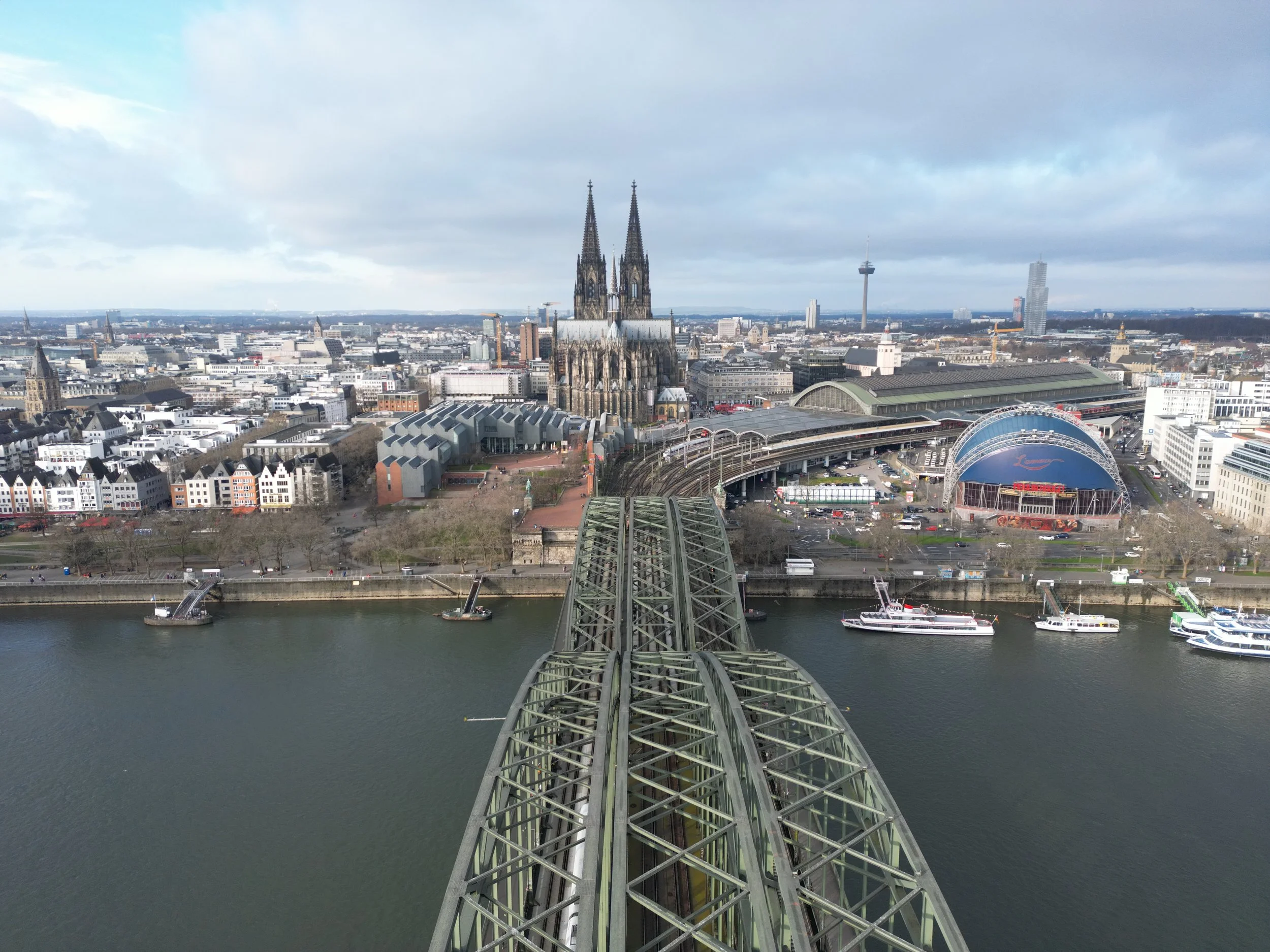 Aerial view of Cologne, Germany, showing the Hohenzollern Bridge over the Rhine River with boats, the Cologne Cathedral with twin spires, and cityscape including a telecommunications tower and modern buildings under a cloudy sky.