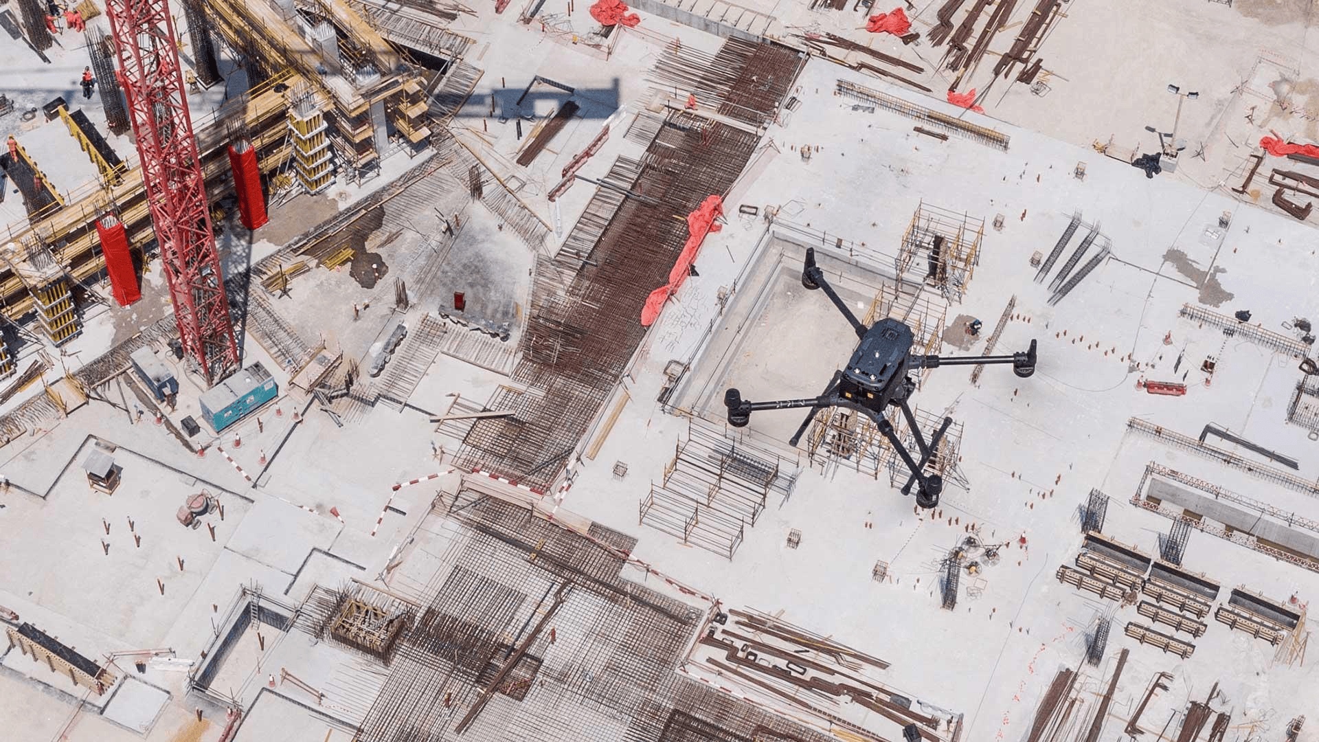 An aerial view of a construction site with a red crane, concrete foundation, and steel reinforcements, surrounded by scaffolding and construction workers.