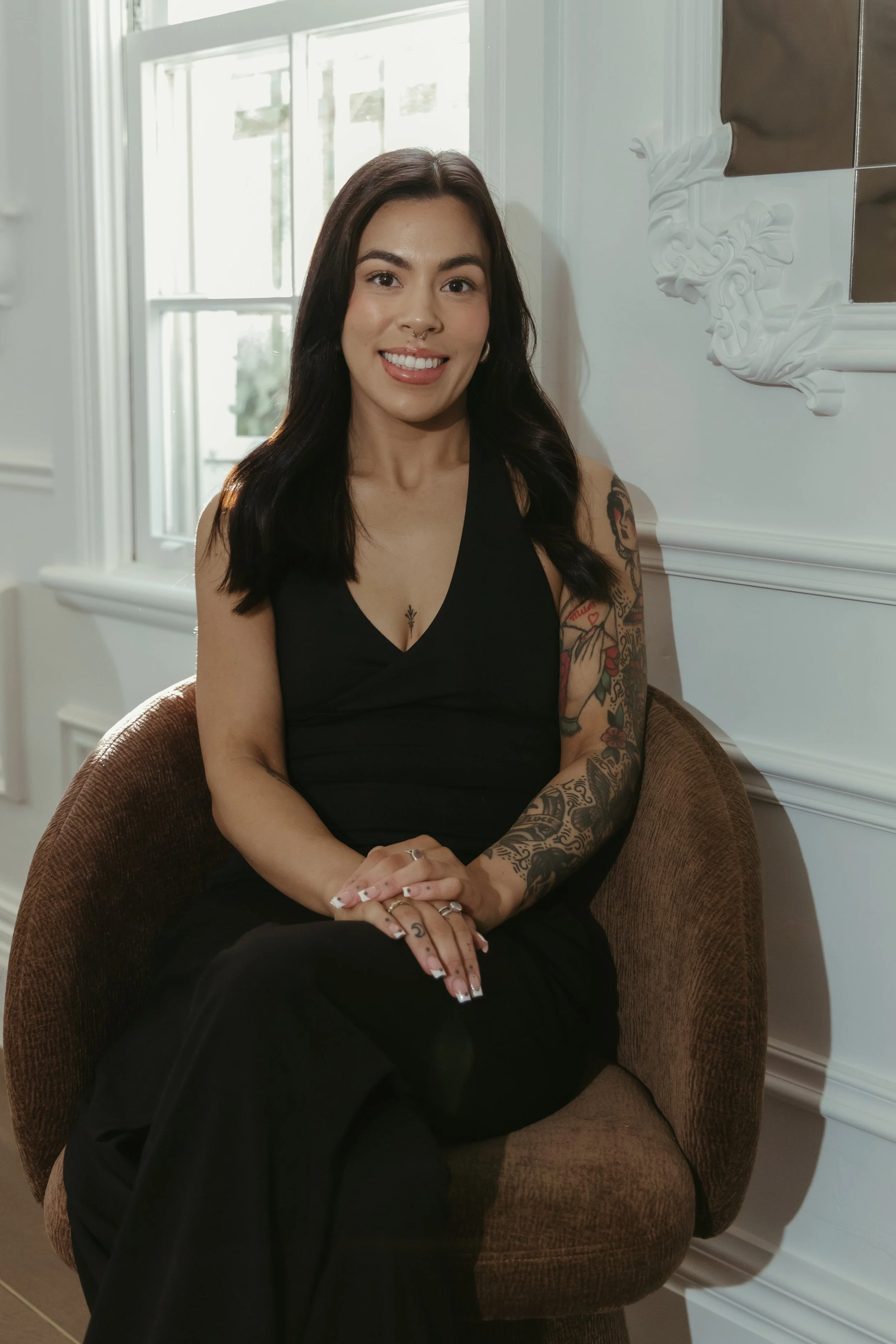 A smiling woman with long dark hair and tattoos on her arm, sitting in a brown armchair, wearing a black sleeveless top, in a well-lit room with white walls, decorative molding, and a window with blinds.