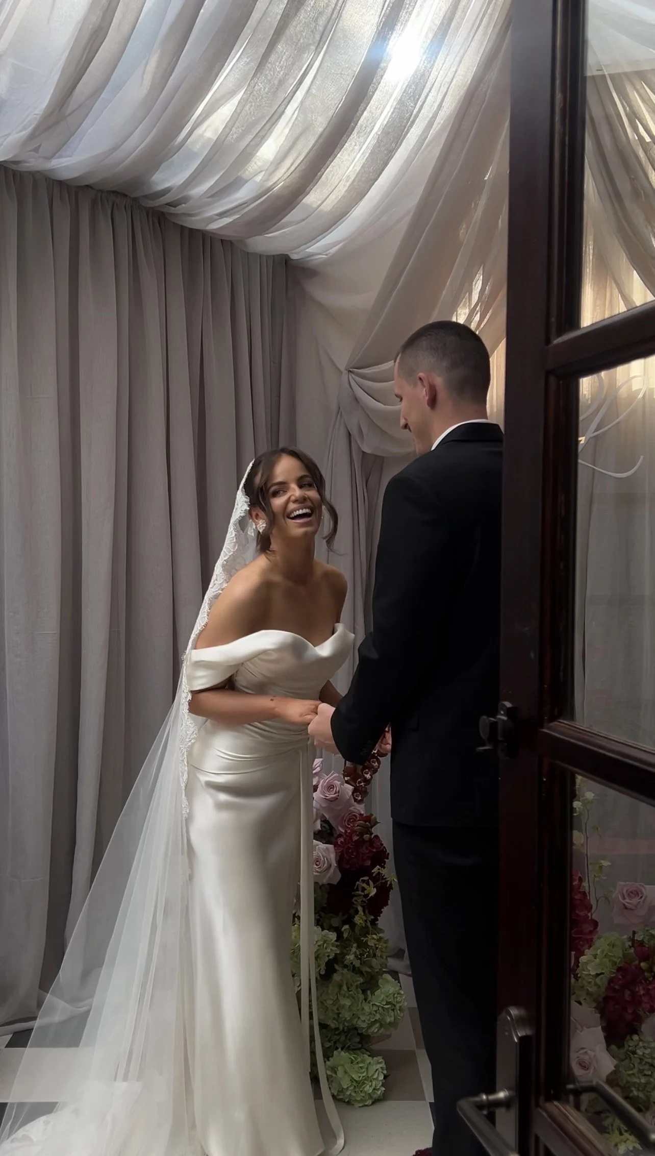 A bride and groom are holding hands and smiling during their wedding ceremony, standing in front of a curtain backdrop with flowers at their feet.