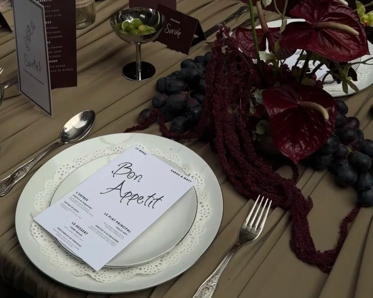 Table setting with a white dinner plate, a menu card with French text, a silver fork and spoon, and a centerpiece of red and purple flowers, grapes, and greenery on a wooden table.