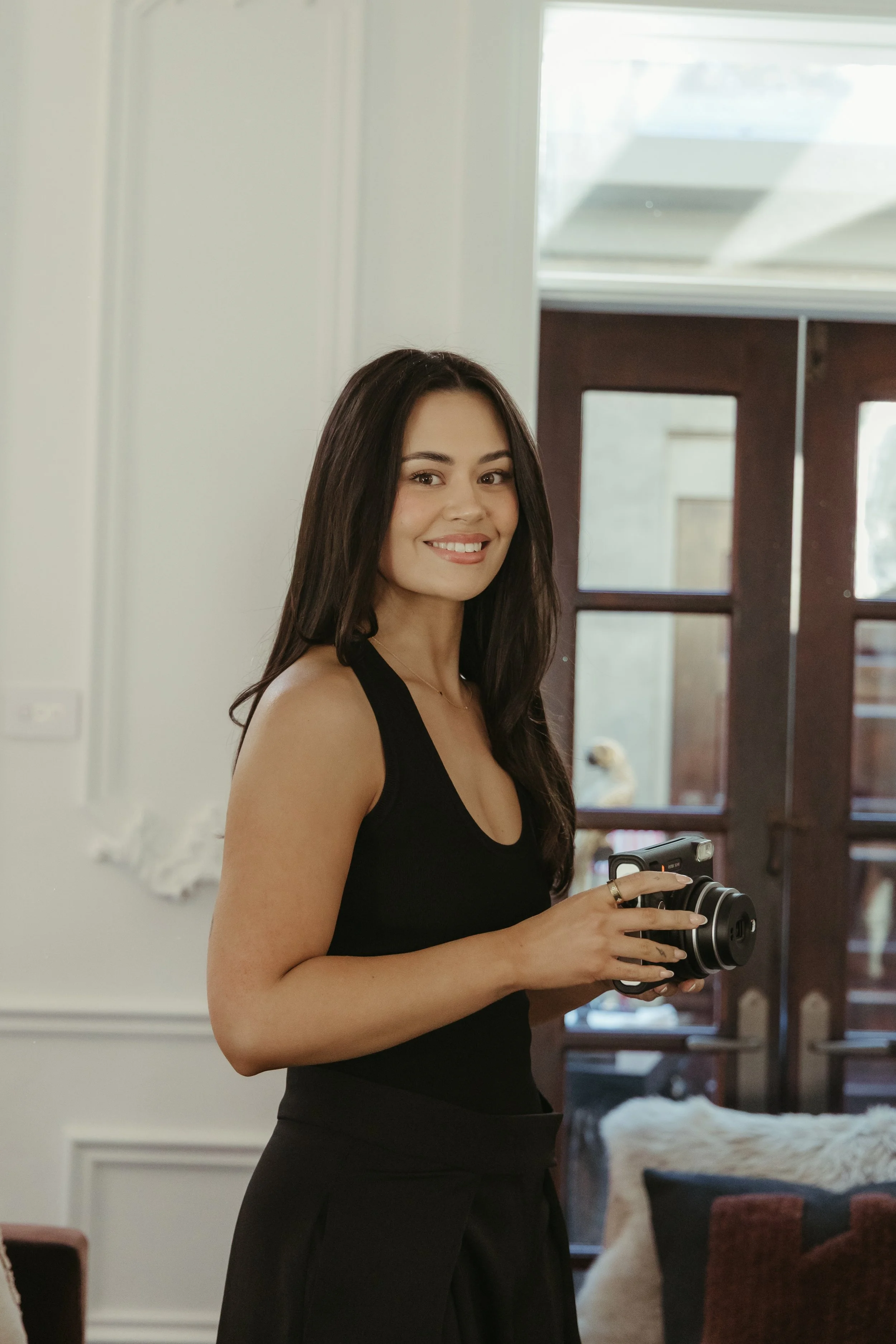 A smiling woman with long dark hair wearing a black sleeveless top and black skirt holding a camera in her hands standing indoors near a door with glass panels and a window, with a dog and furniture visible in the background.