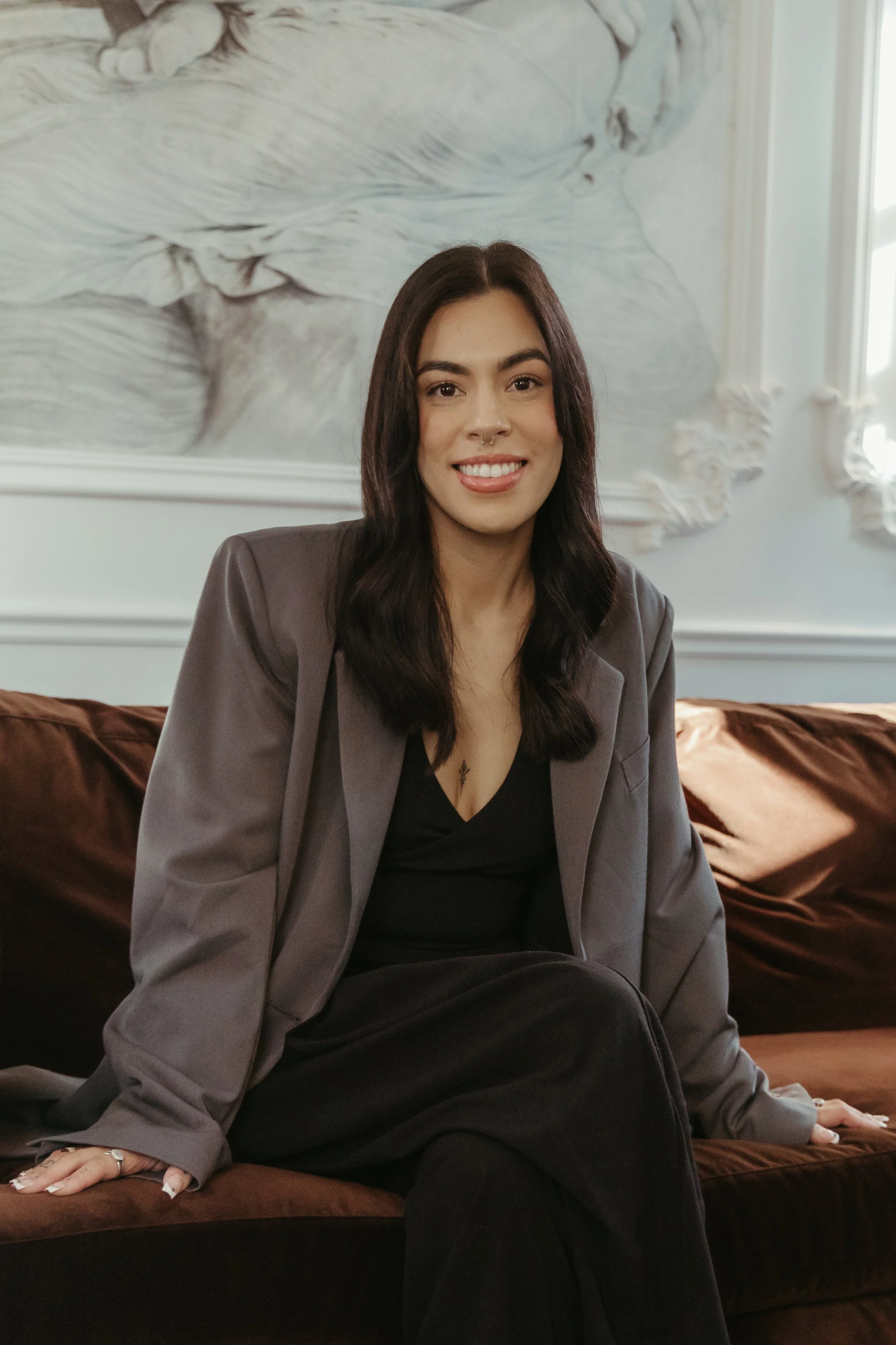 A woman with long dark hair, wearing a gray blazer and black top, sitting on a brown sofa with a marble relief sculpture in the background.