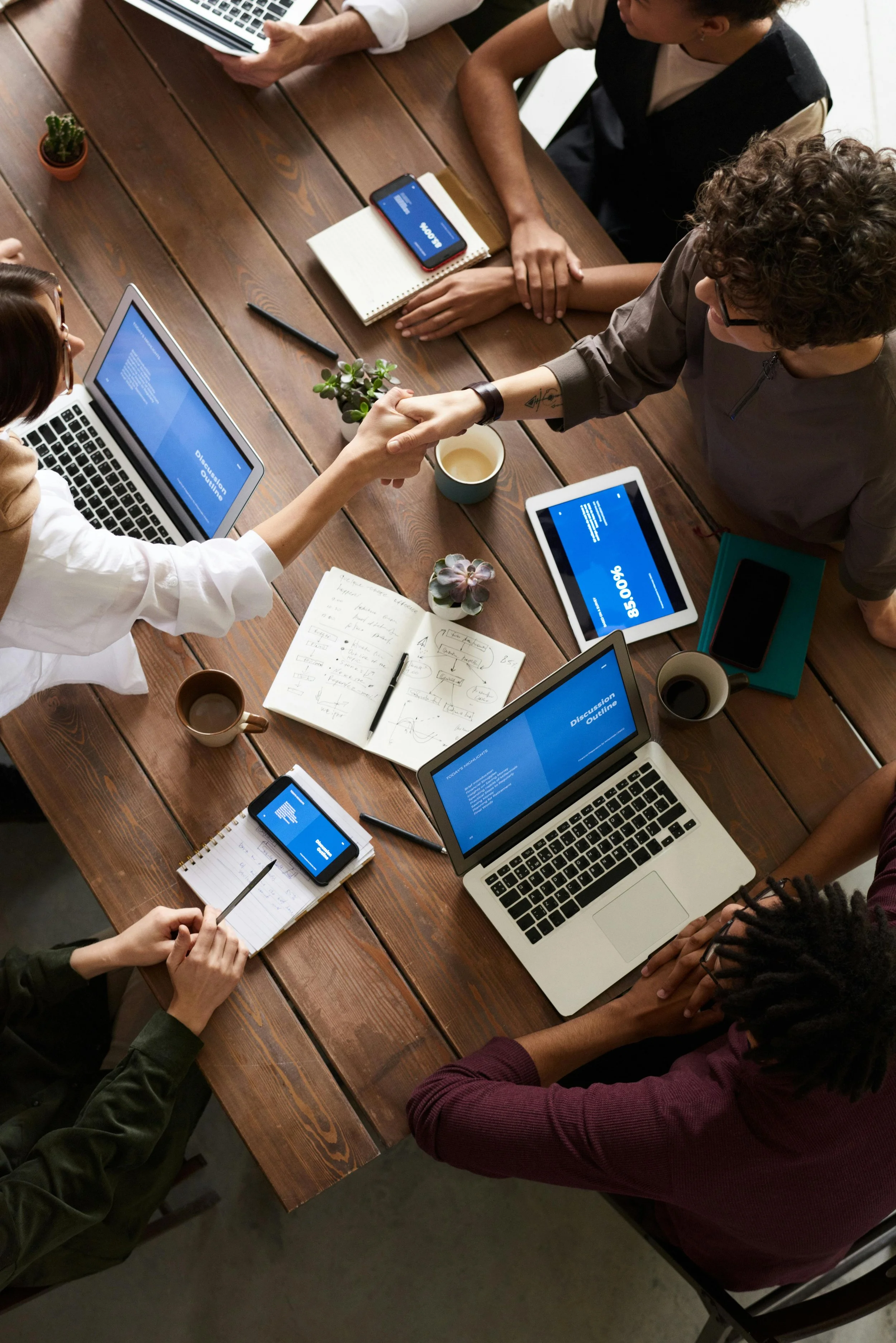 Five people sitting around a wooden table with laptops, tablets, notebooks, and cups, shaking hands in the center, engaging in a meeting or discussion.