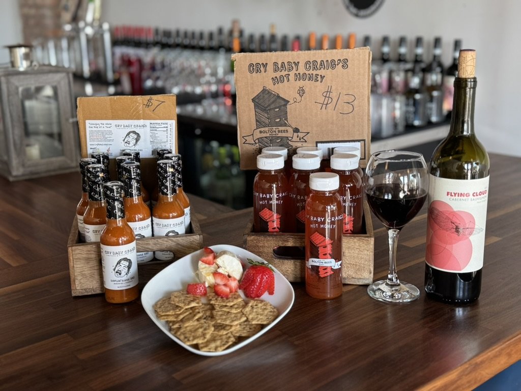 Display of hot honey sauces, a glass of red wine, and a bottle of cabernet wine on a wooden counter, with a plate of crackers and strawberries in the foreground.